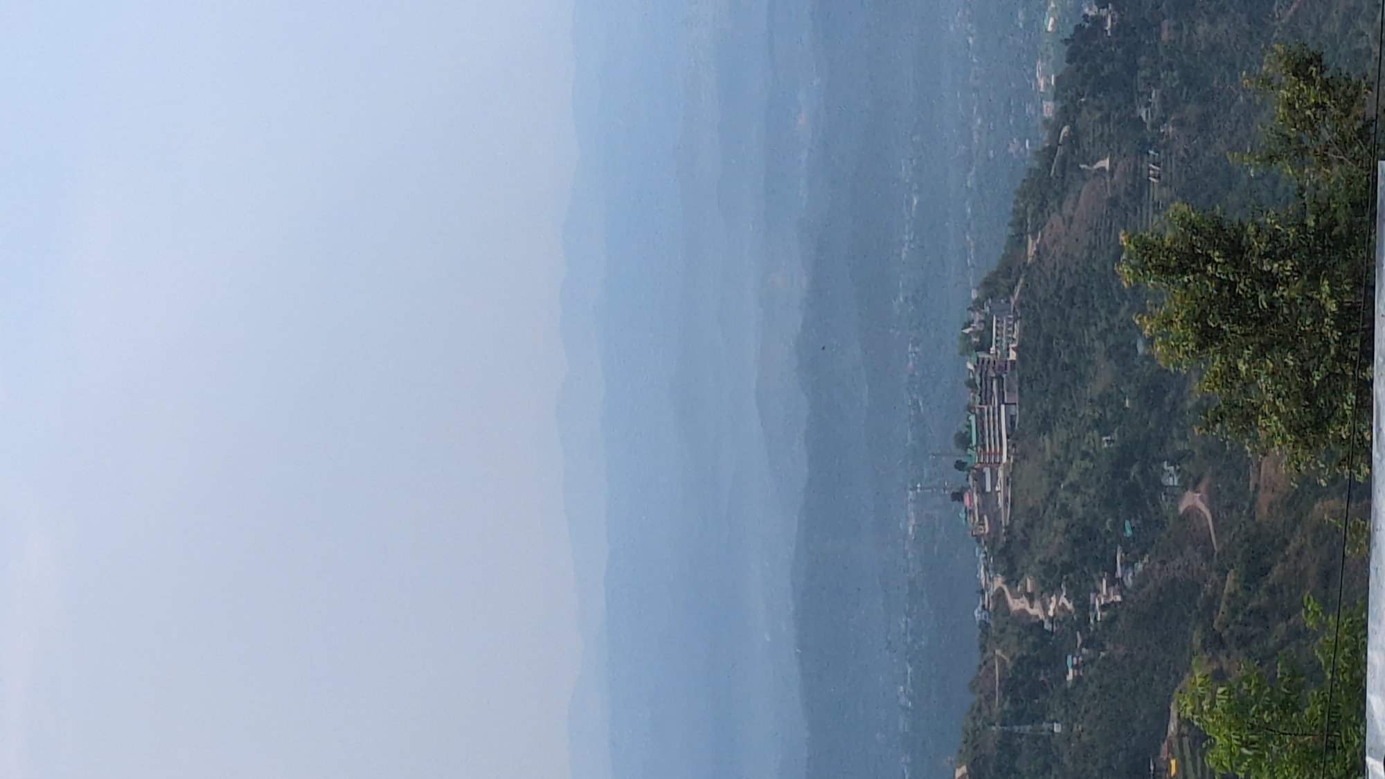 The image displays a vast mountainous landscape, captured from a high vantage point, with the original orientation appearing to be a panoramic horizontal view presented vertically. In the immediate foreground, on the right side of the frame (if viewed horizontally), dense green foliage and tree branches partially frame the scene. Below these trees, a hillside slopes down, dotted with numerous buildings that form a small settlement or village. These structures, visible with varying roof colors and architectural styles, are nestled on the terraced terrain, suggesting adaptation to the hilly environment. A prominent tall structure, possibly a tower or antenna, stands amidst the buildings. Winding roads or pathways are also discernable on the slopes.
Beyond this foreground, multiple layers of mountains extend into the distance, progressively fading into shades of blue and gray, characteristic of atmospheric haze or mist. The receding peaks create a sense of immense depth and scale. The sky, occupying the left half of the frame (if viewed horizontally), is a clear, light blue, indicating daytime conditions, likely late morning or early afternoon, contributing to the overall hazy appearance of the distant landscape. There are no discernible people or specific activities taking place; the image is a static landscape view. No visible text is present. The scene strongly suggests a remote, hilly, or mountainous region, consistent with the provided location context of Haini, India.