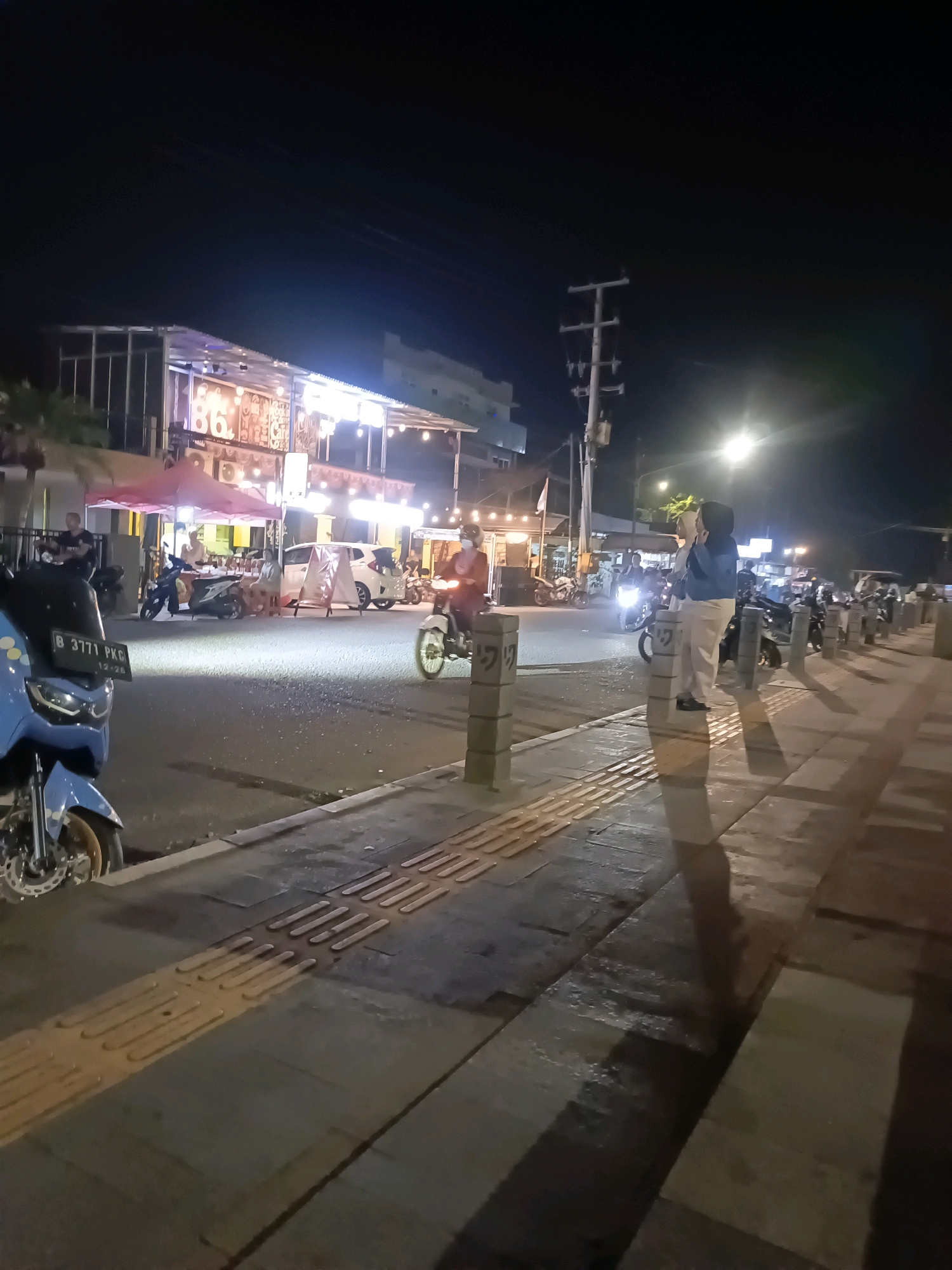 This image captures a nocturnal street scene in Lubuklinggau, Indonesia, likely a commercial or entertainment area given the presence of illuminated food stalls or restaurants. A bright blue motorcycle is prominently parked in the foreground, with its license plate readable as "B 3771 PKG". Across the street, an establishment with "86" displayed on its facade is lit up, with patrons seated under a red awning. Several other motorcycles are visible, some in motion, with riders wearing helmets, suggesting ongoing traffic. Two individuals, a woman in a hijab and light-colored trousers, and another person beside her, are standing on a sidewalk lined with tactile paving. The sidewalk is bordered by concrete bollards with decorative markings. The street itself appears wet, possibly from recent rain, reflecting the ambient light. A utility pole with electrical transformers stands on the right side of the frame, indicating a developed urban setting. The overall atmosphere is lively and active, characteristic of a night market or a street with numerous food and entertainment venues.
