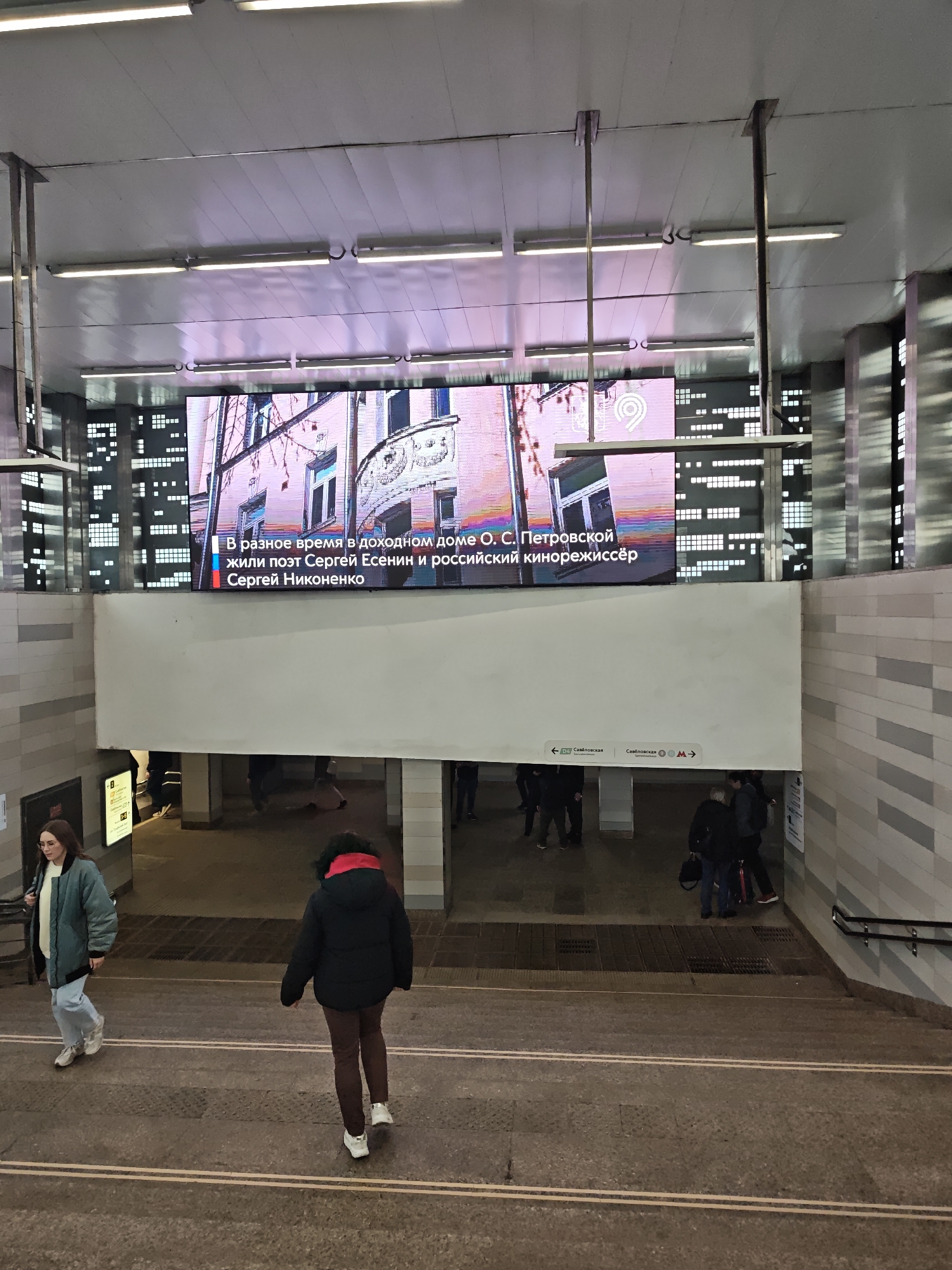 The image captures a public transportation area, likely a subway station in Moscow, Russia. Overhead, a large electronic billboard displays information. The text visible on the screen mentions poet Sergei Yesenin and Russian film director Sergei Nikonenko, indicating the station may be related to these cultural figures. Signage in Cyrillic script reads "Savelovskaya," identifying the station name. People are seen walking up and down the stairs, with two individuals being closer to the camera. The environment is characterized by tiled walls and stairways.