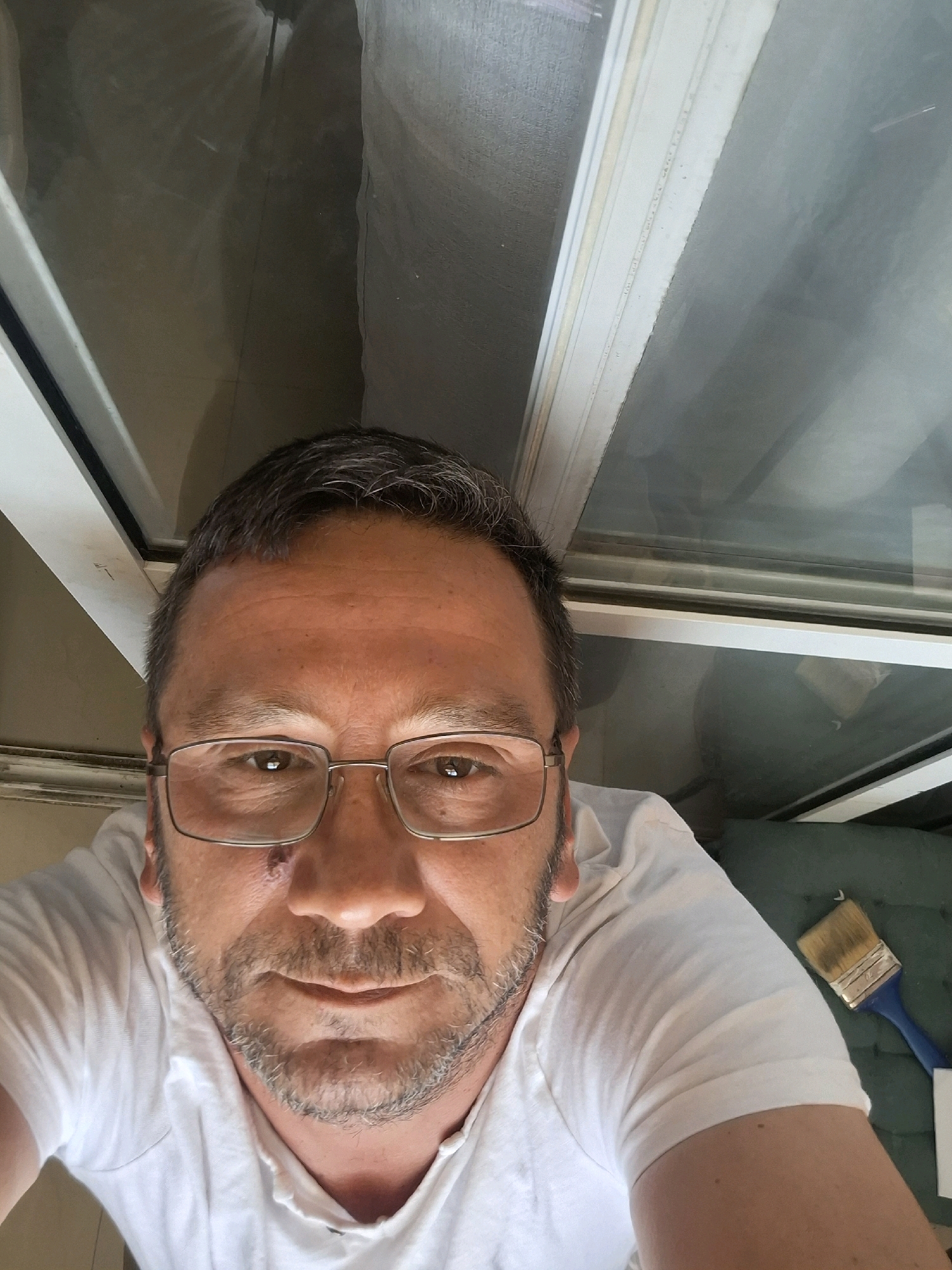 A man with short, graying hair and stubble is taking a selfie. He is wearing a white t-shirt and glasses. A small wound is visible on his cheek. A paintbrush with a blue handle is lying on a green surface to his right. The background appears to be indoors, with large windows reflecting a gray sky and what looks like a portion of a building. The image is shot from a high angle, looking down at the man.