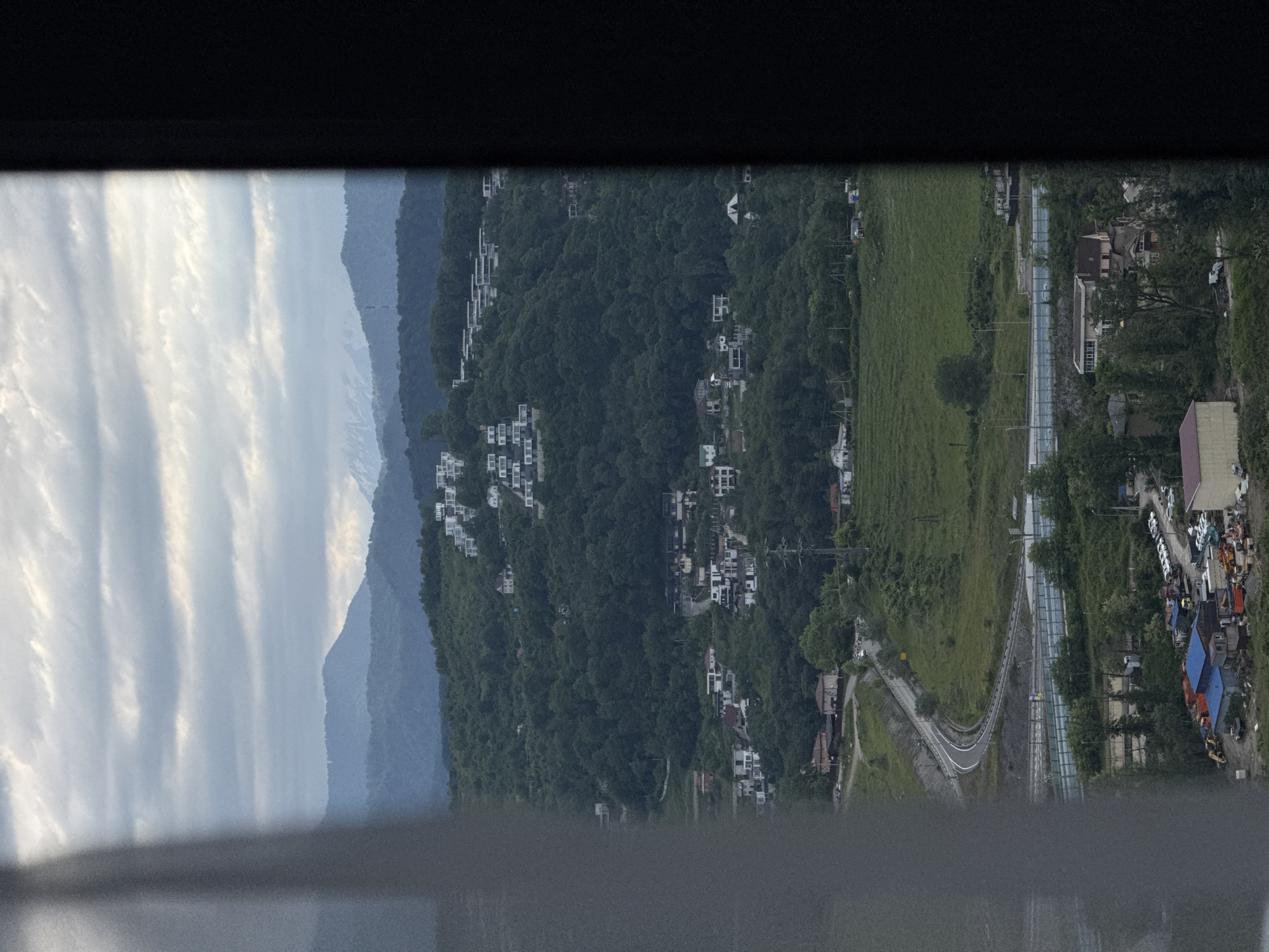 The image captures a panoramic view from an elevated position in Sochi, Russia. The left side of the frame shows a hazy mountain range under a sky with striated clouds. Progressing rightward, a densely forested hillside is dotted with white residential structures. This transitions into a green field adjacent to a roadway that curves through the landscape. In the lower-right portion, there is evidence of human activity, with a line of parked vehicles and structures covered in blue tarpaulins.