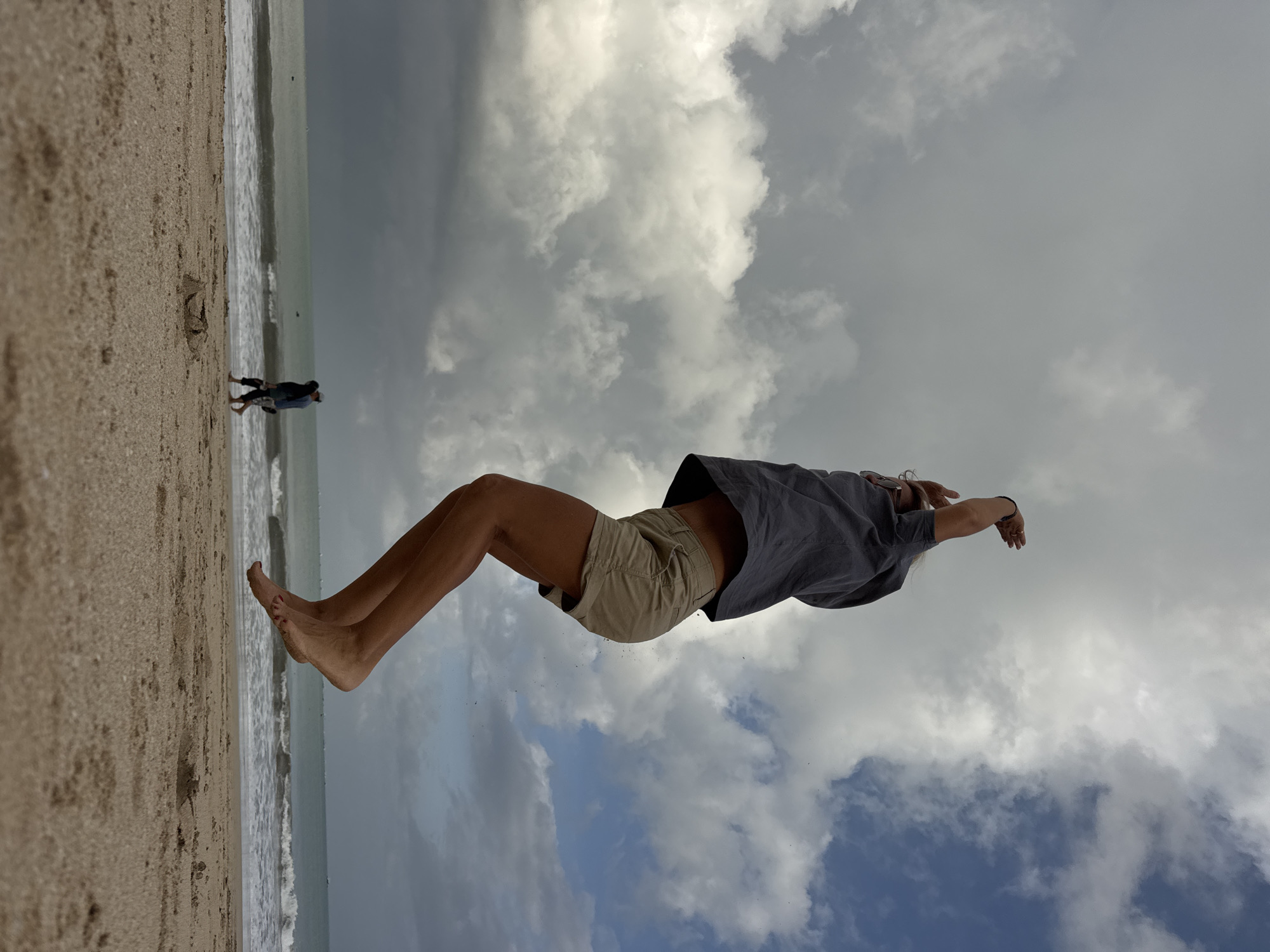 A low-angle, medium shot captures a person mid-jump on a sandy beach. The subject, wearing a grey short-sleeved shirt and beige shorts, is suspended in the air with their arms raised above their head. Their legs are bent, and their feet are pointed downwards, suggesting an upward leap. The background features a beach with gentle waves rolling onto the shore, and a cloudy sky with patches of blue. In the distance, two people are walking along the shoreline, and several small objects, possibly birds, are scattered across the sky. The scene appears to be the Kuta beach in Bali, Indonesia.