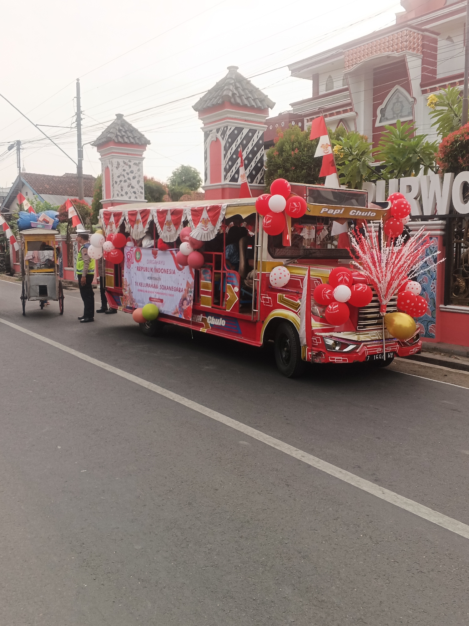 The image shows a red and yellow decorated vehicle, possibly a small bus or a modified truck, adorned with red and white balloons and flags. The vehicle has a banner that reads "DIRGAHAYU REPUBLIK INDONESIA" and "TK KELURAHAN SOKANEGARA". Two individuals in uniform, one appearing to be a traffic officer, stand next to the vehicle on a paved road. A small cart is visible to the left of the officer. In the background, there are buildings with red and white architectural details, suggesting a festive or official event. The environment appears to be an outdoor setting in a city. The license plate of the vehicle is visible and reads "B 1604 HY".