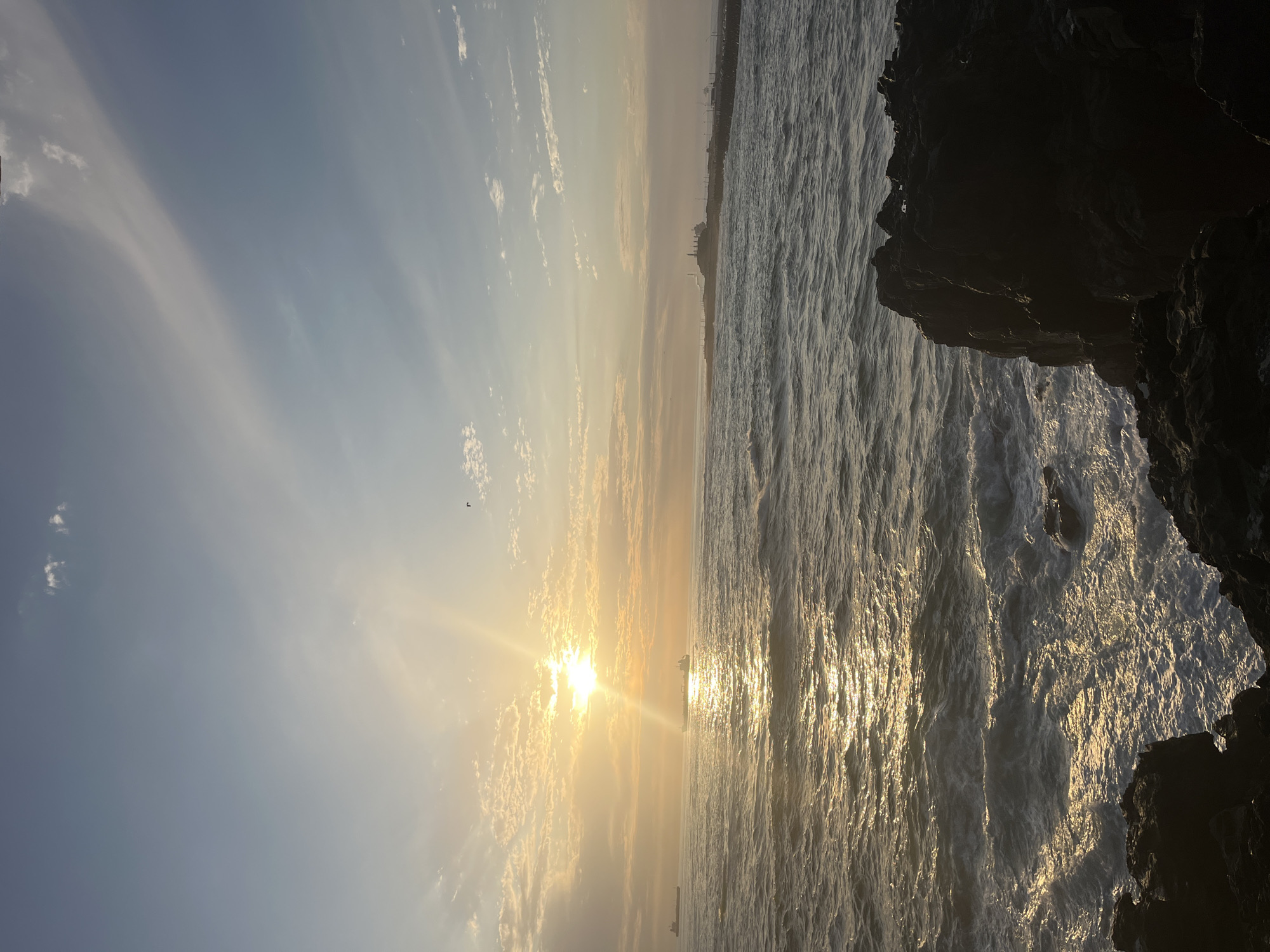 The image depicts a coastal scene at sunset, likely taken in Arica, Chile, based on the city skyline visible on the horizon. The sun is positioned low in the sky, creating a bright glare and reflecting off the ocean surface, resulting in a shimmering golden path across the water. The sky is a mix of blue and yellow hues, with scattered clouds. To the right of the image, there is a dark, rocky cliff formation. Waves are actively breaking on the shore. There is a bird and at least two boats visible in the distance.