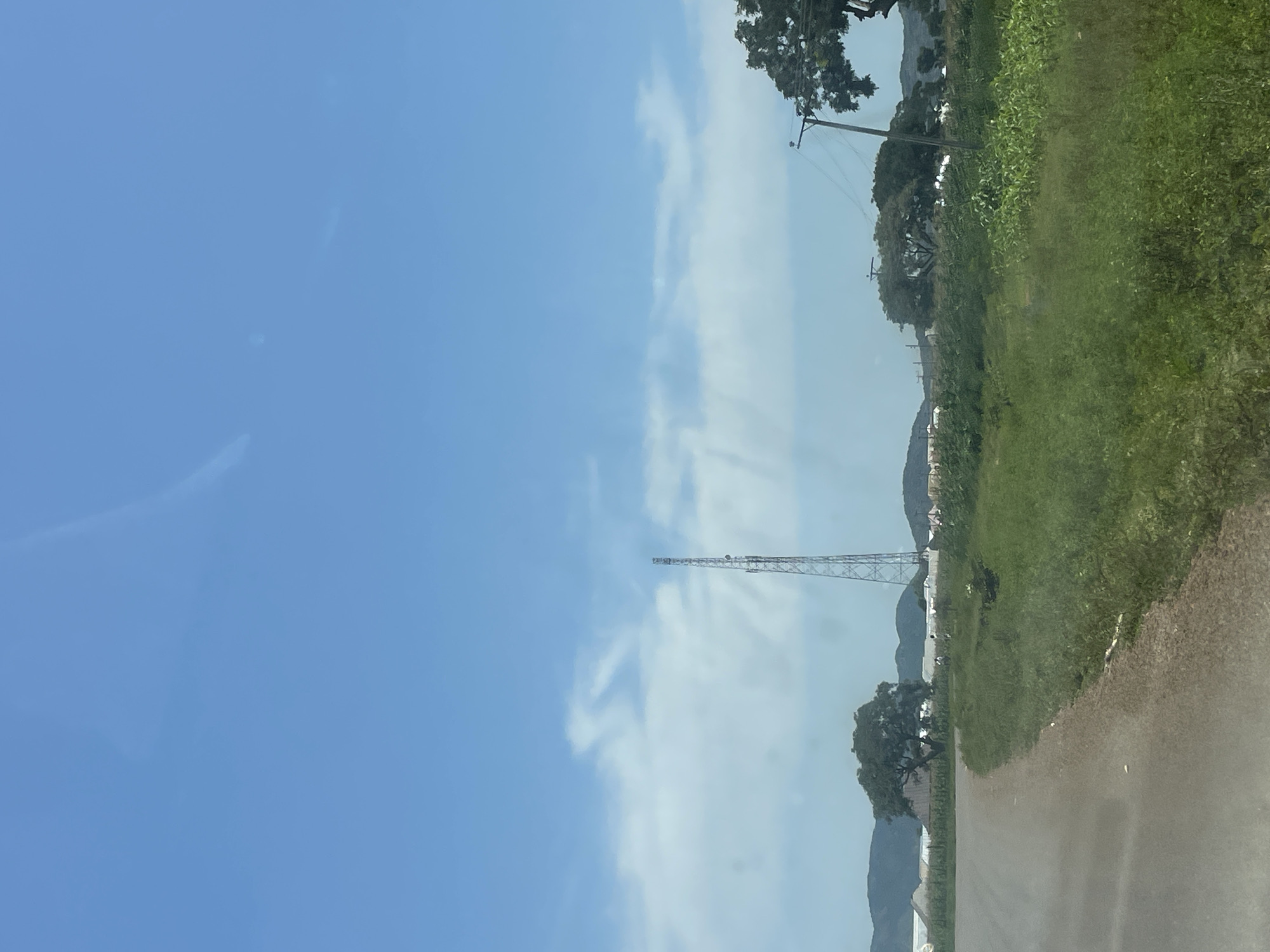 The image depicts a rural landscape under a bright, partly cloudy sky. A tall, metallic communication tower stands prominently in the middle ground. In the foreground, a paved road curves to the left, bordered by a grassy verge. The background features rolling hills, several trees, and some structures that appear to be greenhouses or agricultural buildings. A wooden utility pole with wires is visible on the right side of the image. The scene is serene and appears to be set in an open, undeveloped area, likely outside of a densely populated urban center. The location is identified as Zalau, Nigeria.
