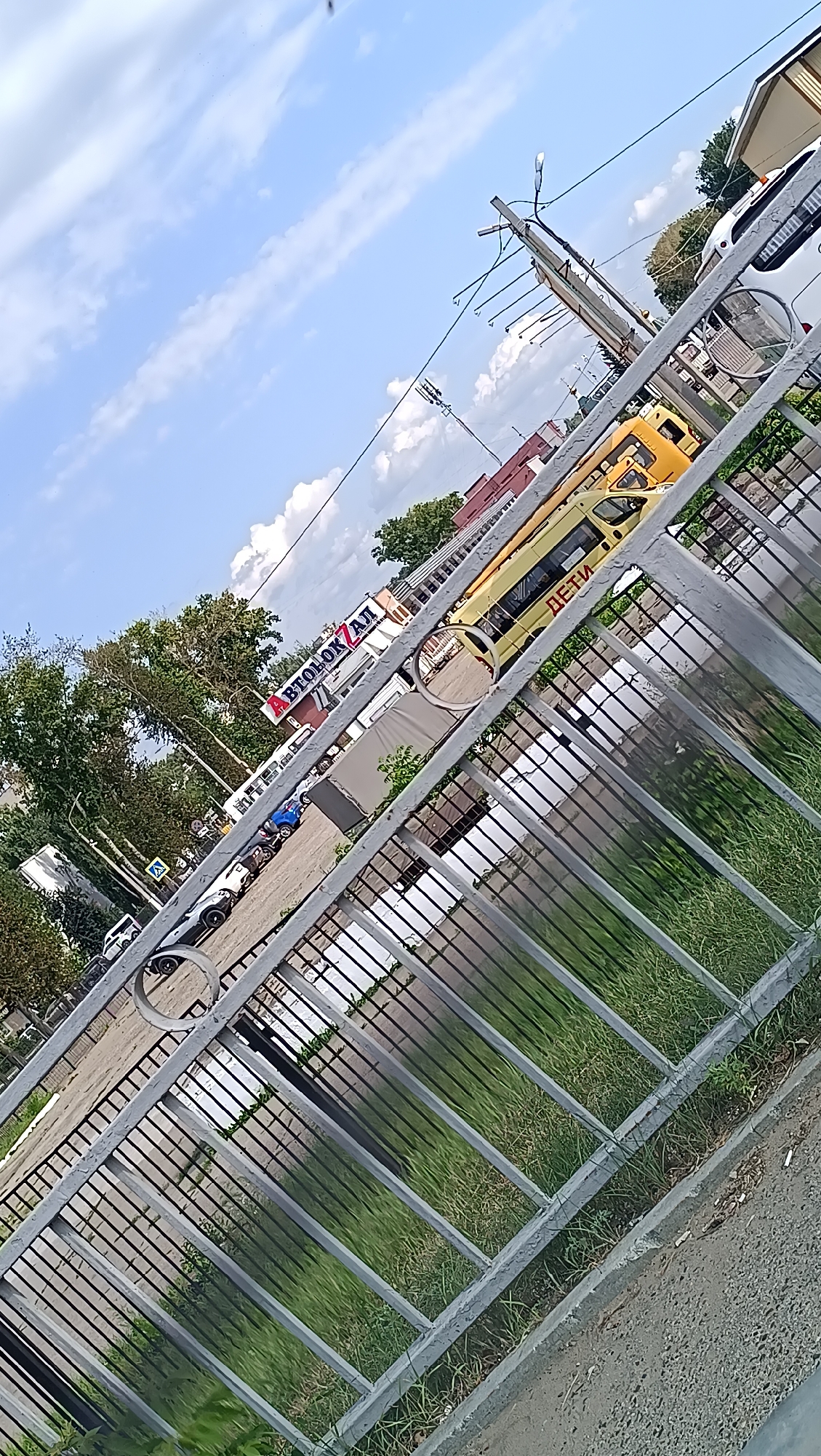 The image, taken in Chaikovsky, Russia, shows a street scene from behind a grey metal fence. In the background, there is a yellow bus with the Cyrillic word "ДЕТИ" (children) written on its side. Next to it, a white van or truck is parked, and further down the street, a building with a sign displaying Cyrillic text, possibly "ВТОЛОК АЛ", is visible. Several cars are parked along the side of the road. Trees line the street, and the sky is blue with scattered white clouds. The foreground is dominated by the fence and green grass growing on the ground. The image is taken at an angle, suggesting the photographer is looking downwards or from an elevated position.