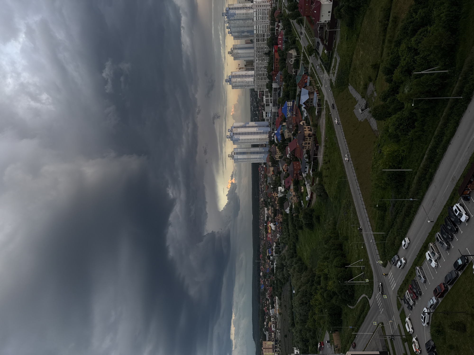 This is a high-angle, wide-shot aerial photograph taken in the city of Perm, Russia, likely during a period of stormy weather as indicated by the dark, heavy clouds dominating the sky. The scene captures a suburban or semi-urban landscape characterized by a mix of residential buildings and green spaces.
In the foreground, a multi-lane road network is visible, with several cars parked in a lot and others driving on the road. The vehicles are of various makes and models, suggesting a typical urban environment. A few individuals are seen on foot near the roads and parking areas.
Beyond the immediate road network, the landscape transitions into a verdant area with a dense forest or park. Further in the mid-ground, a cluster of multi-story residential buildings, appearing to be apartment complexes, stand out against the natural landscape. These buildings are modern in design, with light-colored facades and blue accents. Adjacent to these larger buildings are numerous detached houses with pitched roofs, indicating a mix of housing types.
The background features a more distant expanse of greenery, possibly extending into the countryside or foothills. The sky is a dramatic display of dark, ominous storm clouds, with some lighter, warmer tones visible on the horizon, possibly from the sun attempting to break through or reflecting off distant clouds. The overall lighting suggests a dim, overcast day. The image appears to have been taken from a high vantage point, possibly a tall building or drone, offering a comprehensive view of the urban and natural elements.
