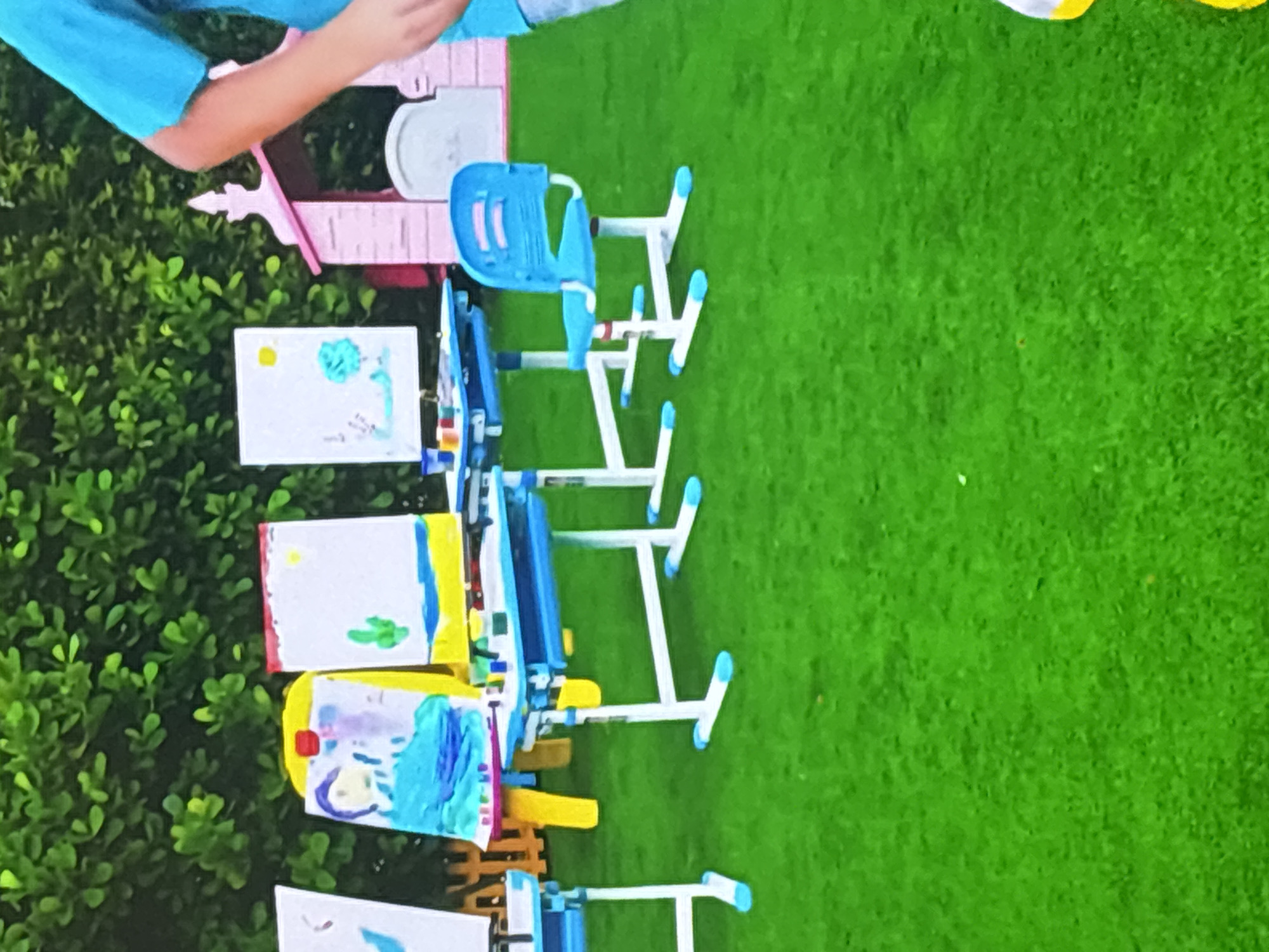 The image depicts an outdoor art setup on a vibrant green lawn, likely artificial turf, with lush green foliage in the background. In the foreground, several children's art desks with blue and white frames are arranged. Each desk has a white canvas placed on an easel. One canvas displays a drawing of a person in a blue dress and a yellow sun. Another canvas features a drawing of a cactus and a yellow sun. A third canvas shows a light blue tree under a yellow sun. On the desks, there are cups of paint and brushes. A bright blue child-sized chair is positioned next to one of the desks. In the background, a pink playhouse with a white roof and arched windows is visible. To the right of the frame, a person wearing a bright blue t-shirt is partially visible, suggesting participation in the art activity. The scene appears to be a children's art class or an outdoor creative space. Given the bright lighting and the presence of green foliage, the image could be from a park, a backyard, or an educational facility with outdoor space in Dubai, United Arab Emirates.