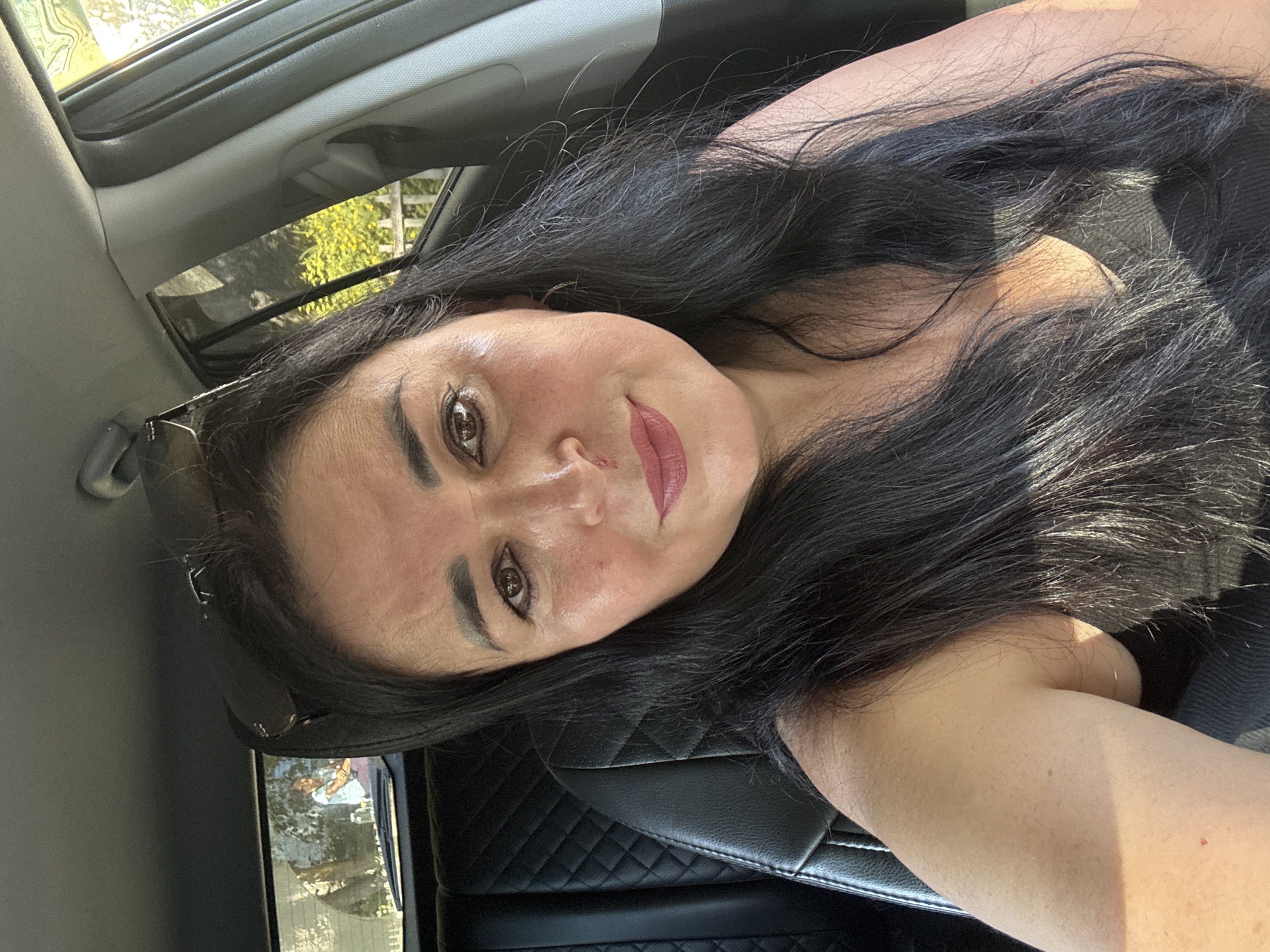 The image shows a woman sitting in the driver's seat of a car. She has long, dark hair and is wearing a black top. Her face is partially visible, and she is looking towards the camera with a slight smile. Sunglasses are perched on top of her head. The interior of the car is visible, including the black leather seat and part of the dashboard. Through the rear window, two people can be seen in the backseat, with one appearing to be a child. Outside the car, trees and a fence are visible, suggesting a suburban or natural environment. The lighting suggests it is daytime, possibly late afternoon due to the warm sunlight. The specific location in Novotroitsk, Russia, cannot be determined from the visual information provided in the image.
