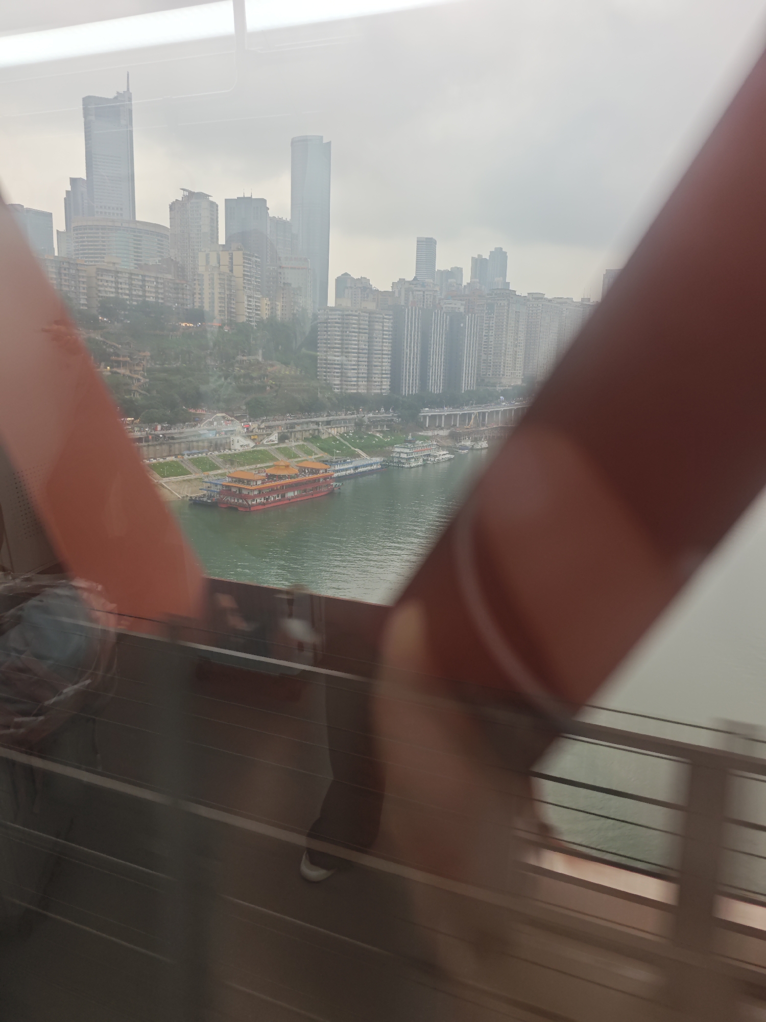 The photo, taken from inside a moving train or vehicle on a bridge, shows a partial view of the Yuzhong District in Chongqing, China.
A blurry, red structural support of the bridge dominates the right and left sides of the frame. The cityscape is visible in the background: a mix of high-rise buildings and apartment complexes. A river flows in the mid-ground, with several boats docked near the shore. The weather appears overcast or hazy, muting the colors.
Inside the vehicle, a person's leg and foot are visible, suggesting the photo was taken by a passenger. A metal railing is also within view, indicating the interior of the train or vehicle.