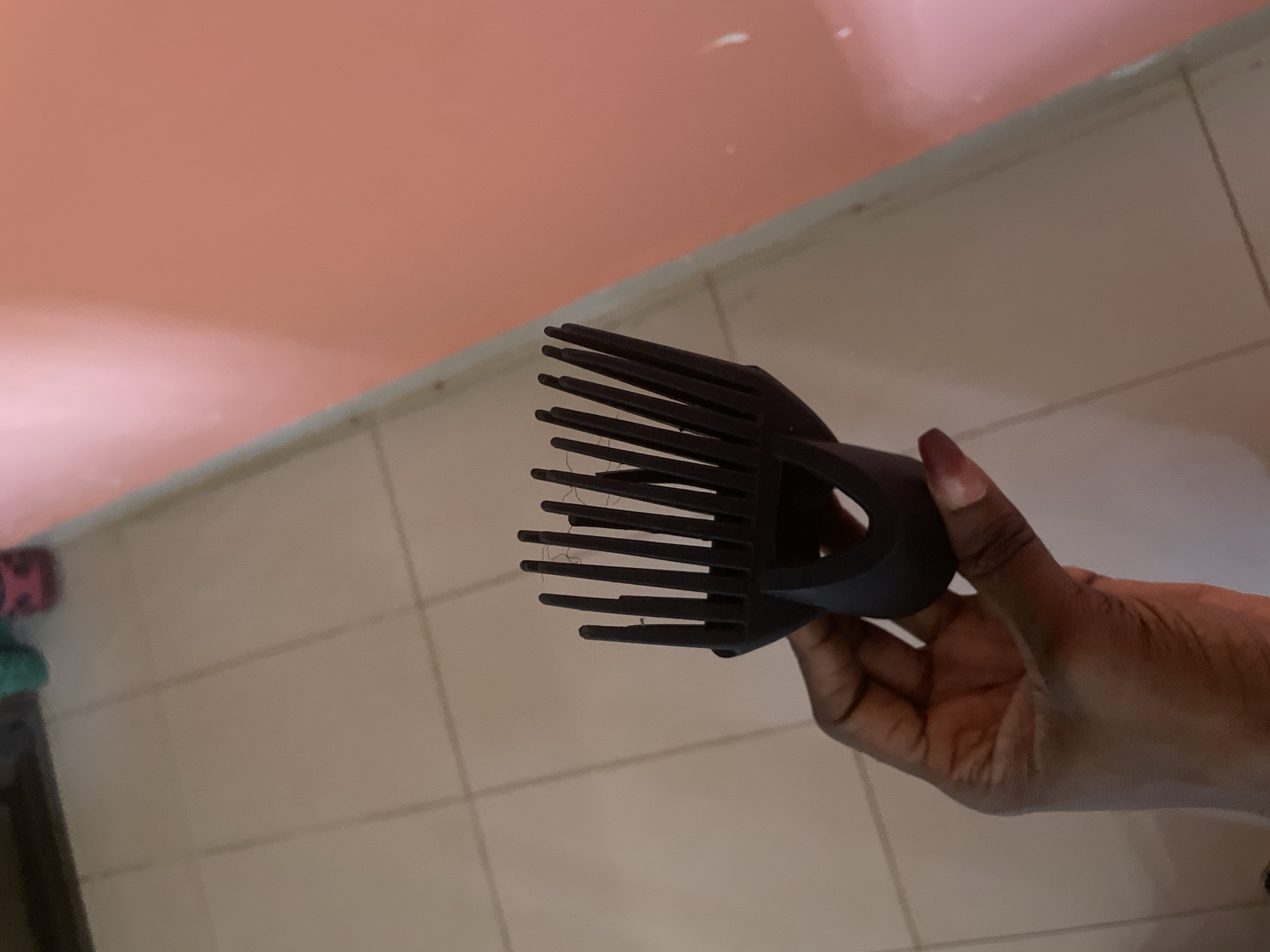 A person's hand, exhibiting dark skin tone and manicured fingernails, holds a black diffuser attachment for a hairdryer. The attachment features a wide comb-like structure with numerous prongs, some of which appear to have dark strands of hair caught between them. The background consists of tiled flooring with a light beige hue and a pink-colored wall. The scene is indoors, suggesting a domestic environment. This photograph was taken in Kakau, Nigeria.