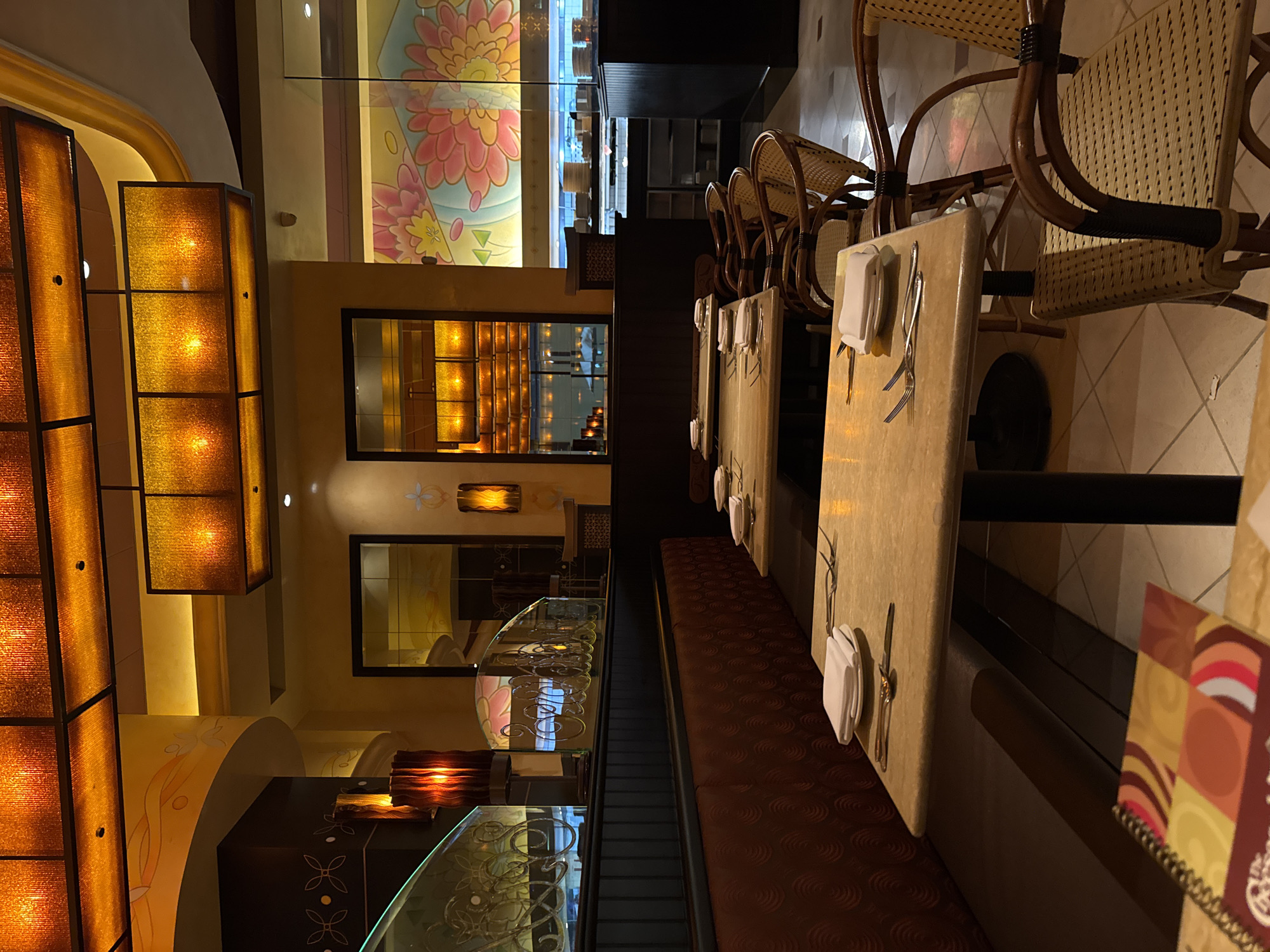 The image shows the interior of a restaurant with tables set for dining. The tables are long and narrow, with light-colored surfaces and place settings including napkins and silverware. A bench with dark red upholstery runs along one side of the tables. Wicker chairs are placed opposite the bench. The lighting is warm and subdued, with illuminated decorative panels on the walls. A glass partition with floral designs is visible in the background. The scene suggests a restaurant setting, possibly in a Cheesecake Factory location. The city of Puebla, Mexico, is not readily apparent from the visual information alone. The image does not feature any visible people or actions.
