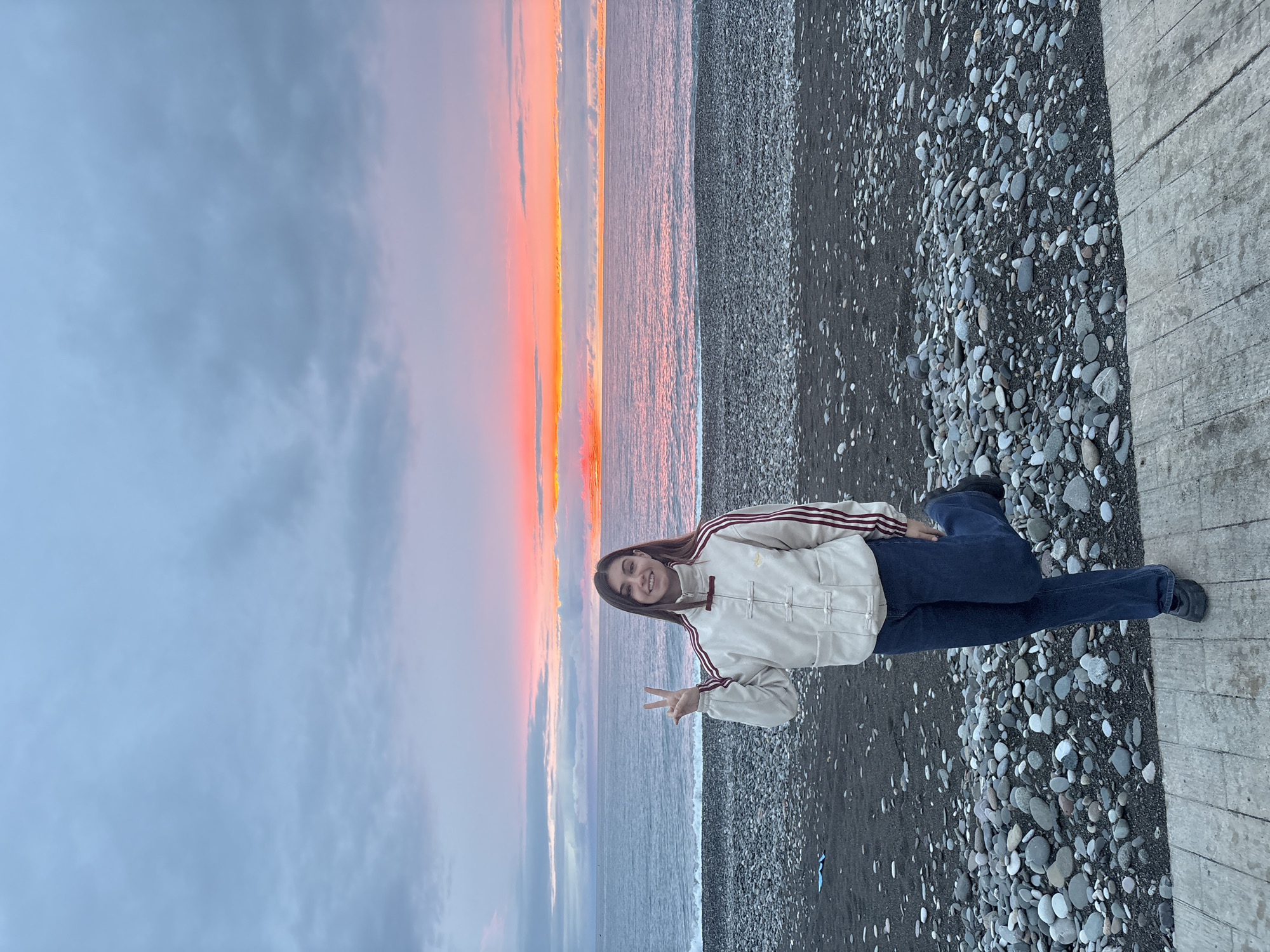 A portrait-oriented photograph captures a woman posing at what appears to be a beach in Batumi, Georgia, during a sunset. The woman has long, dark hair, wears a light-colored jacket with dark stripes, and blue jeans. She is seated and gives a peace sign to the camera. The environment includes a stone beach and a body of water. In the background, the sky is illuminated by the vibrant hues of a sunset, with the clouds colored in shades of gray and pink.