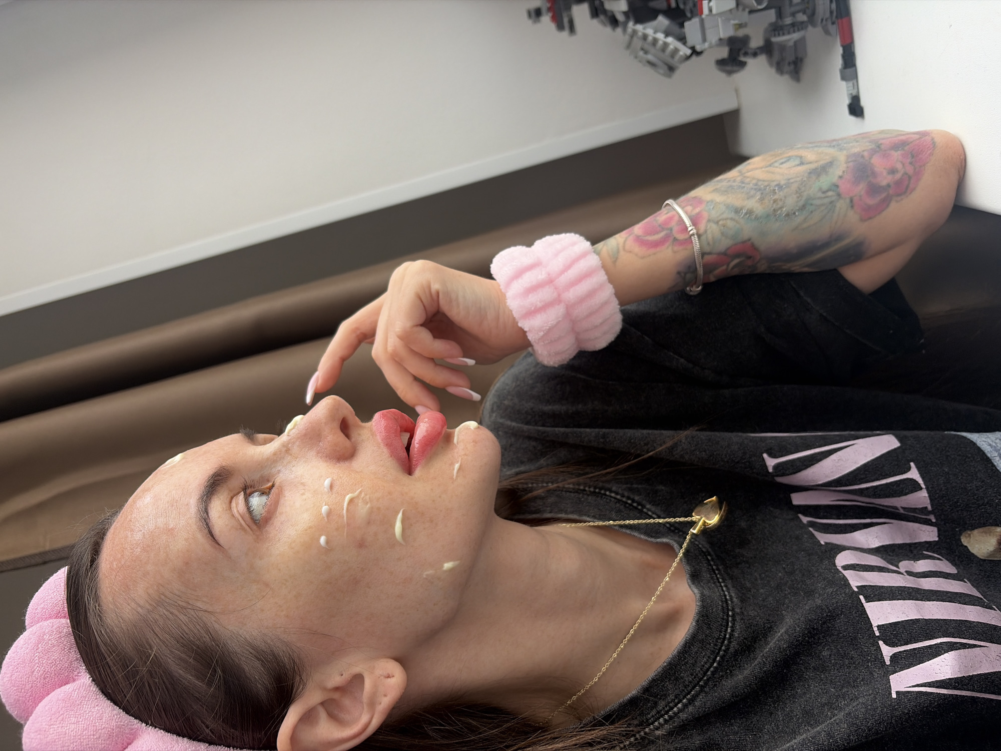 A woman is captured in a close-up, high-angle shot, lying down indoors. She is positioned with her head slightly tilted back and to the left, looking upwards. Her face has freckles and several dots of a light-colored cream, suggesting a skincare routine or mask application. Her lips are slightly parted.
She wears a soft, pink, fluffy headband around her hairline and a matching wristband on her right wrist. A dark, possibly charcoal-colored, long-sleeved top covers her torso, featuring light pink text, a portion of which reads "NIE". A delicate gold chain with a small heart-shaped pendant hangs around her neck, resting on her top. Her left arm, adorned with colorful tattoos primarily featuring floral designs, is bent, and a silver bracelet is visible on her wrist.
The background shows a minimalist indoor setting with a light-colored surface (possibly a wall or table) and a darker, soft surface behind her. In the upper right corner, a complex, gray and black object, possibly a Lego construction, is visible, appearing to be mounted or placed on a shelf. The overall impression is one of relaxation and self-care. The location context provided for the image is Saint Petersburg, Russia.