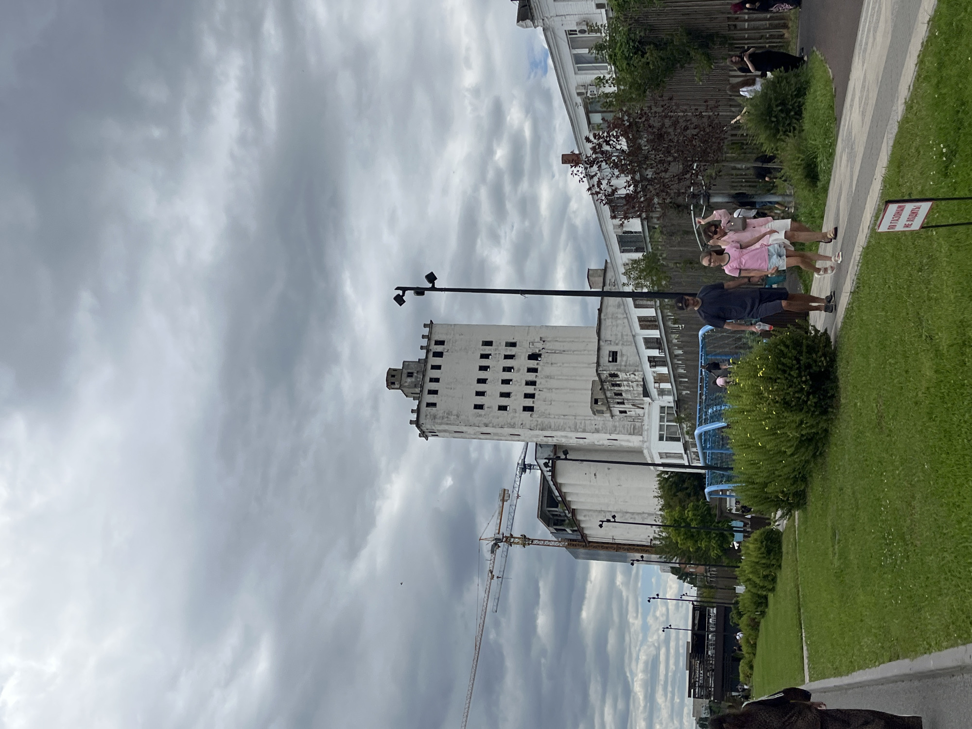 The image depicts an overcast day in Saratov, Russia. In the background, a tall, white, multi-story industrial building with numerous windows stands out. To the left of this building, a construction crane is visible, suggesting ongoing development or renovation. In the mid-ground, a group of people are walking along a paved path. Among them are two women in pink tops and light-colored shorts, and a man in a dark t-shirt and shorts. Further in the background, there is a blue play structure. The foreground consists of a grassy area with manicured bushes. A red and white sign with Russian text is placed on the grass. The sky is heavily clouded, casting a diffused light on the scene.