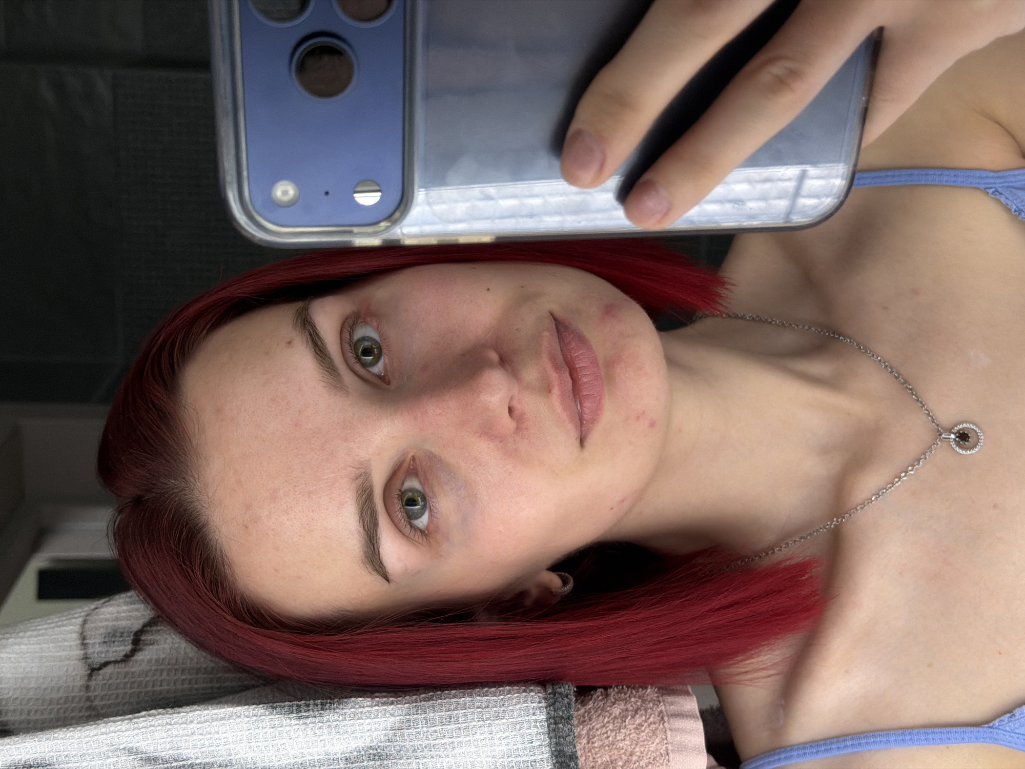The image presents a close-up selfie of a young adult female, taken indoors. The woman is positioned with her head resting on a light-colored, textured towel or fabric, and her body extending towards the right of the frame, giving the impression she is lying down. She has striking red hair, styled down, and clear green eyes that look directly towards the camera. Her complexion is fair, with some minor blemishes visible on her forehead and chin. She wears a light blue top or bra, the strap of which is visible on her right shoulder and chest. A delicate silver chain necklace with a small, circular pendant featuring a dark center stone adorns her neck.
Her left hand holds a smartphone, visible in the upper portion of the frame. The phone, which appears to be a model with multiple large camera lenses (suggesting an iPhone Pro series), is encased in a clear or translucent cover. The reflection of a hand is visible on the phone screen. The background behind her head is dark and indistinct.
The activity depicted is a selfie, with the subject holding the phone herself. Her expression is neutral and calm. There is no visible text in the image. The lighting appears to be artificial and even, typical of an indoor setting. The image lacks specific location cues for Chaikovsky, Russia, beyond the subject herself.