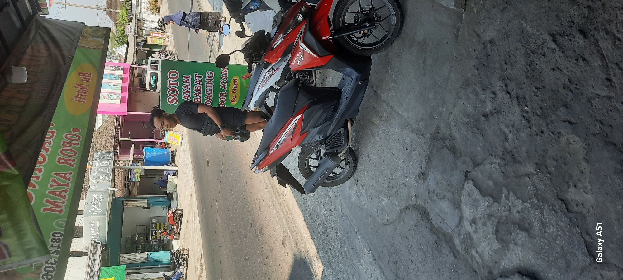 The image depicts a street scene in Selopuro, Indonesia, characterized by a busy commercial area. In the foreground, two motorcycles are parked on a paved road surface that shows signs of wear and tear, with patches of exposed concrete and dirt. To the right, a red Honda motorcycle is prominently featured.
In the mid-ground, a man with short dark hair, wearing a black t-shirt, black shorts, and a dark crossbody bag, stands near a green sign advertising "SOTO AYAM BABAT DAGING POR AYAM Bu Narti." He is looking towards the right of the frame. Behind him, a woman wearing a blue hijab and a patterned dress is walking away from the camera. Several other businesses are visible, including an "Apotek selopuro farma" (pharmacy) with blue accents, and other shops with awnings and signage. A white van is parked further down the street.
The background reveals a slightly hazy sky and the rooftops of buildings, suggesting a typical urban or semi-urban environment in Indonesia. The overall atmosphere suggests a day with bright sunlight, casting shadows on the ground. The signs in the background, though partially obscured, indicate a commercial district. The presence of multiple motorcycles and pedestrians implies daily activity and commerce. The text on the green sign is in Indonesian, identifying the food offered and the proprietor.