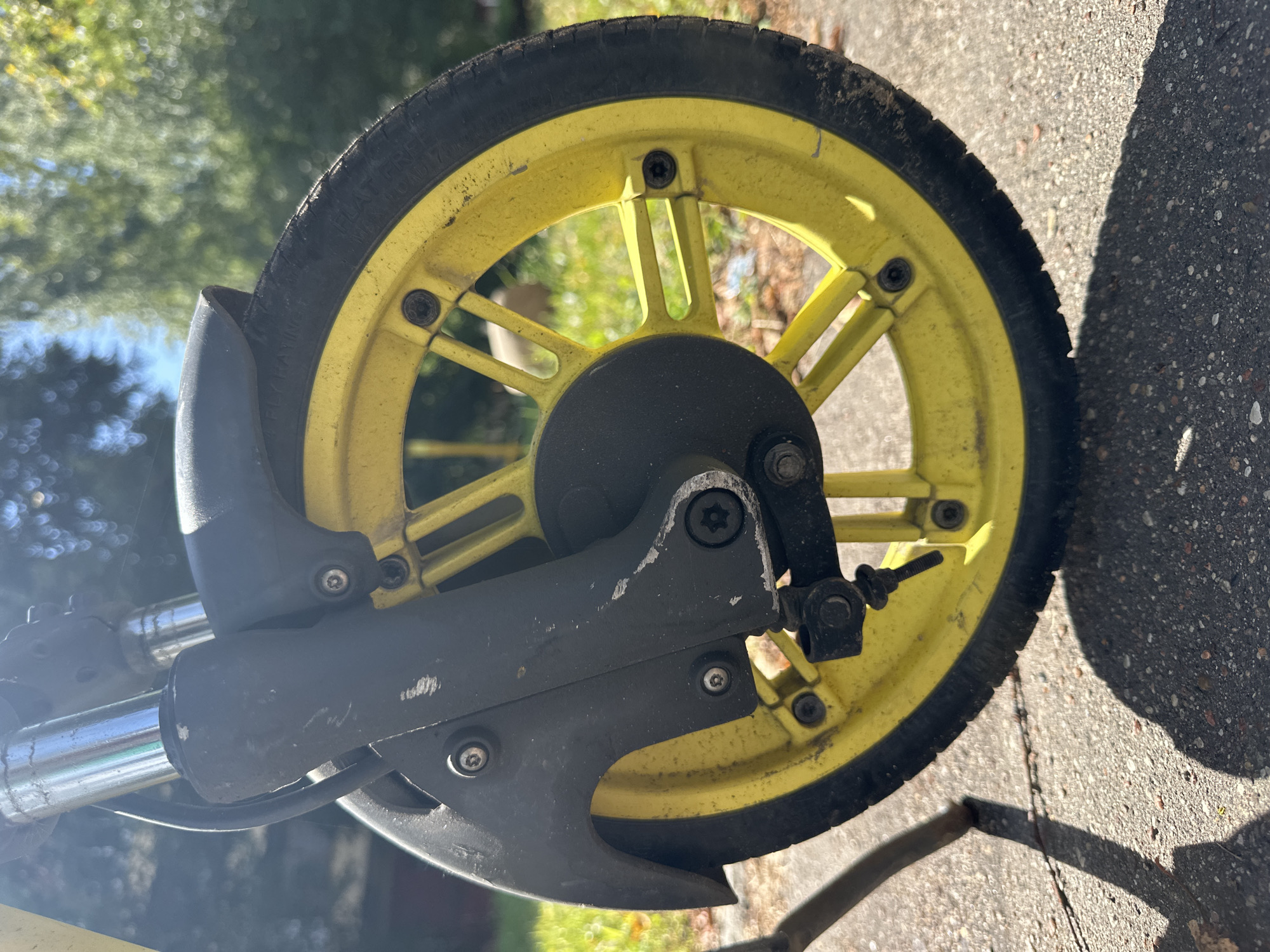 This is a close-up, low-angle outdoor shot of the front wheel and suspension of a yellow and black scooter. The scooter is parked on a grey, speckled asphalt surface, with fallen leaves and debris scattered around. The background shows out-of-focus greenery, suggesting a park or residential area. The scooter's wheel is yellow with black spokes and a black tire. The front fork and fender are black and appear worn and scratched. Sunlight casts a distinct shadow of the scooter's kickstand onto the asphalt in the foreground. The scene does not contain any people or identifiable landmarks suggesting a specific location within Minsk, Belarus.