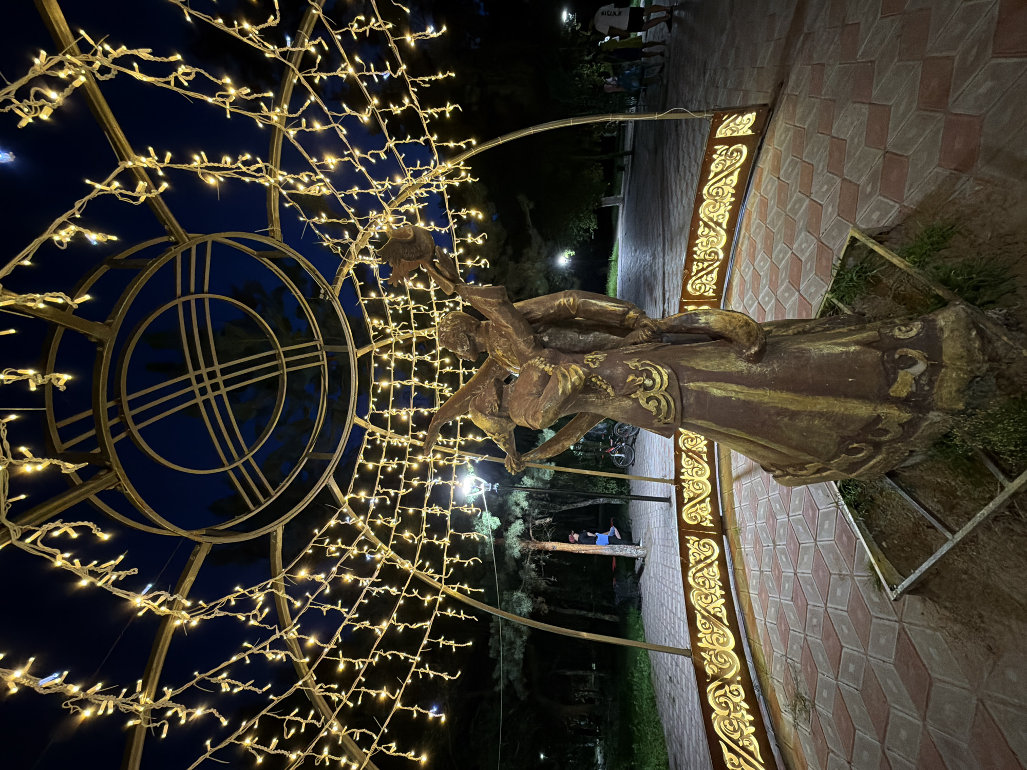 The image captures a bronze sculpture of two figures in an embrace, set within a decorative metal gazebo adorned with string lights. The gazebo has an ornate, golden railing with intricate patterns. The sculpture and gazebo are positioned on a patterned brick pathway in what appears to be a park at dusk or night. The background shows trees and other park elements, including lampposts and distant figures of people. One person is visible on a swing set in the mid-ground, and several people are walking in the background. The overall atmosphere is festive and illuminated by the warm glow of the string lights. This scene is located in the city of Pavlodar, Kazakhstan.