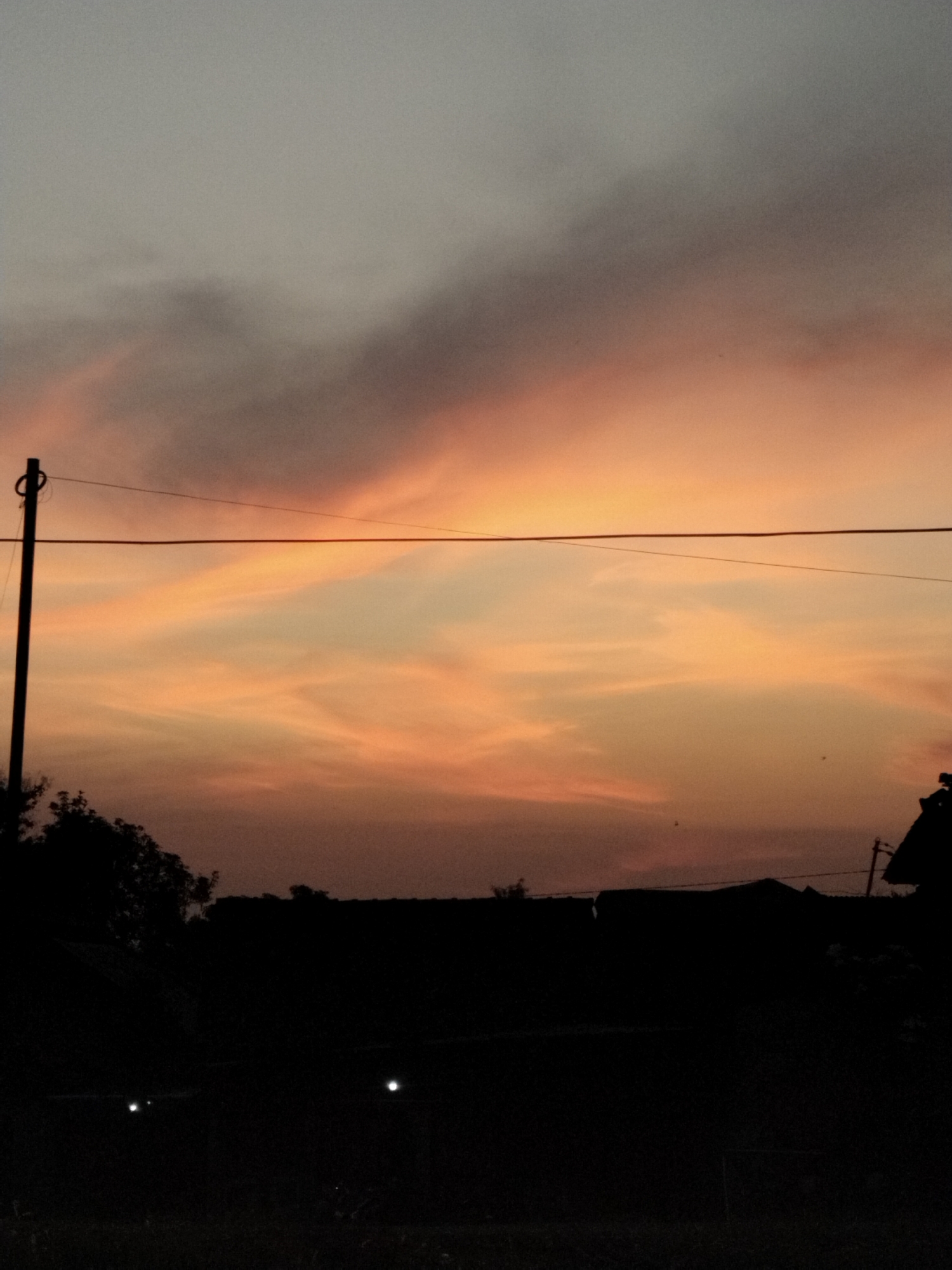 The image captures a silhouetted landscape at dusk. The sky is painted with hues of orange and gray clouds, transitioning into darker tones towards the top. A utility pole with wires stretches horizontally across the mid-section of the frame. Below the sky, the dark silhouettes of trees and rooftops are visible. Two small, bright lights are discernible in the foreground, suggesting artificial illumination. The overall impression is a serene, nocturnal urban or semi-urban scene.