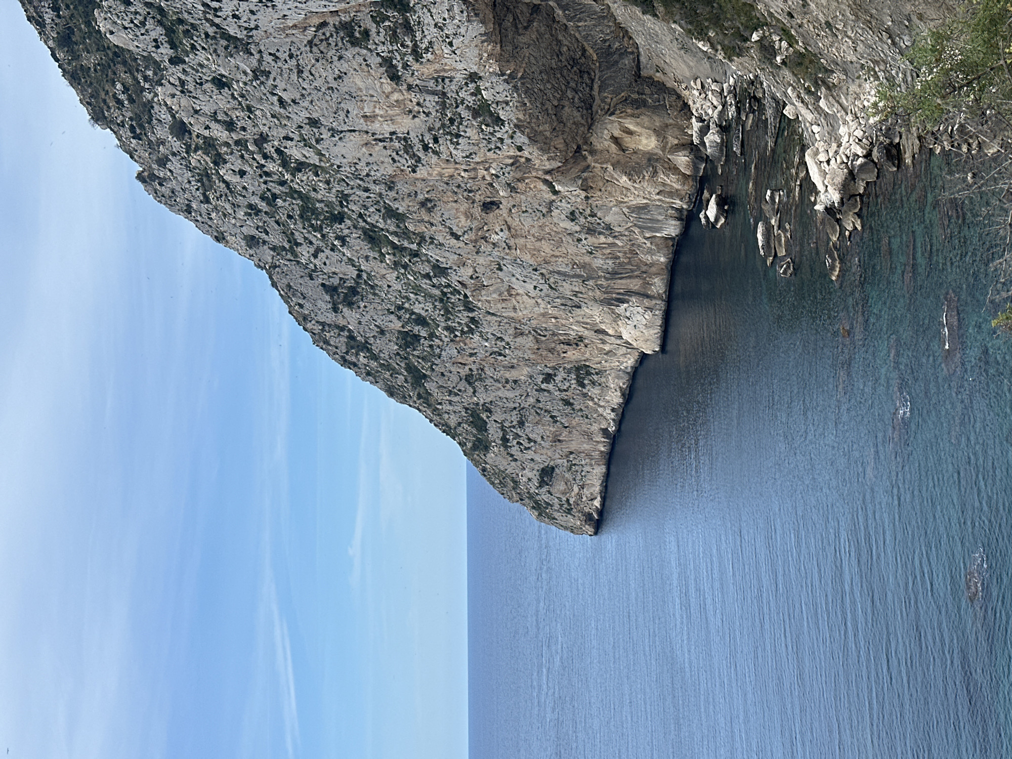 The image shows a coastal scene. A massive rock cliff dominates the frame, dropping sharply into the sea. The rock face is textured and varied in color, from light gray to tan, with patches of dark vegetation. Below the cliff, smaller rocks and boulders line the water's edge, some partially submerged. The sea transitions from a clear, turquoise color near the shore to a deeper blue further out. The sky above is a light, pale blue, seemingly cloudless. The image captures the contrast between the rugged rock formations and the tranquil sea. The location in the city of San Gabriel, Spain, cannot be determined from the image alone.