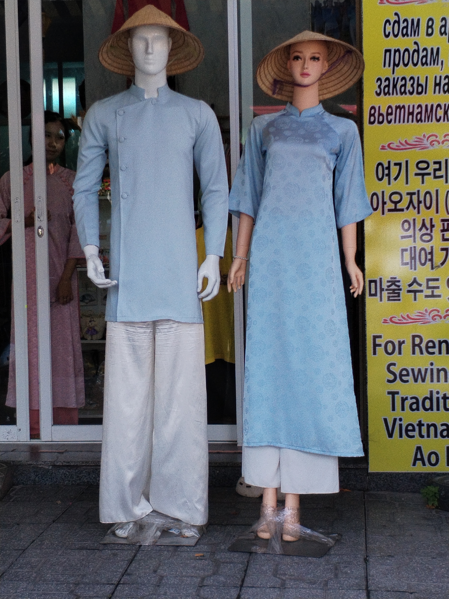 Two mannequins, one male and one female, are displayed outside a storefront in Bac Nha Trang Ward, Vietnam. The male mannequin on the left wears a light blue, long-sleeved tunic with a mandarin collar and seven visible buttons, paired with loose-fitting white trousers. It is topped with a conical straw hat. The female mannequin on the right wears a light blue, knee-length dress with a mandarin collar and decorative textured pattern, also topped with a conical straw hat. Both mannequins have white hands. Behind the mannequins, a person wearing a pink garment is visible through a glass door. A yellow sign with text in Cyrillic, Korean, and English is posted to the right of the mannequins. The mannequins are standing on a grey tiled pavement.