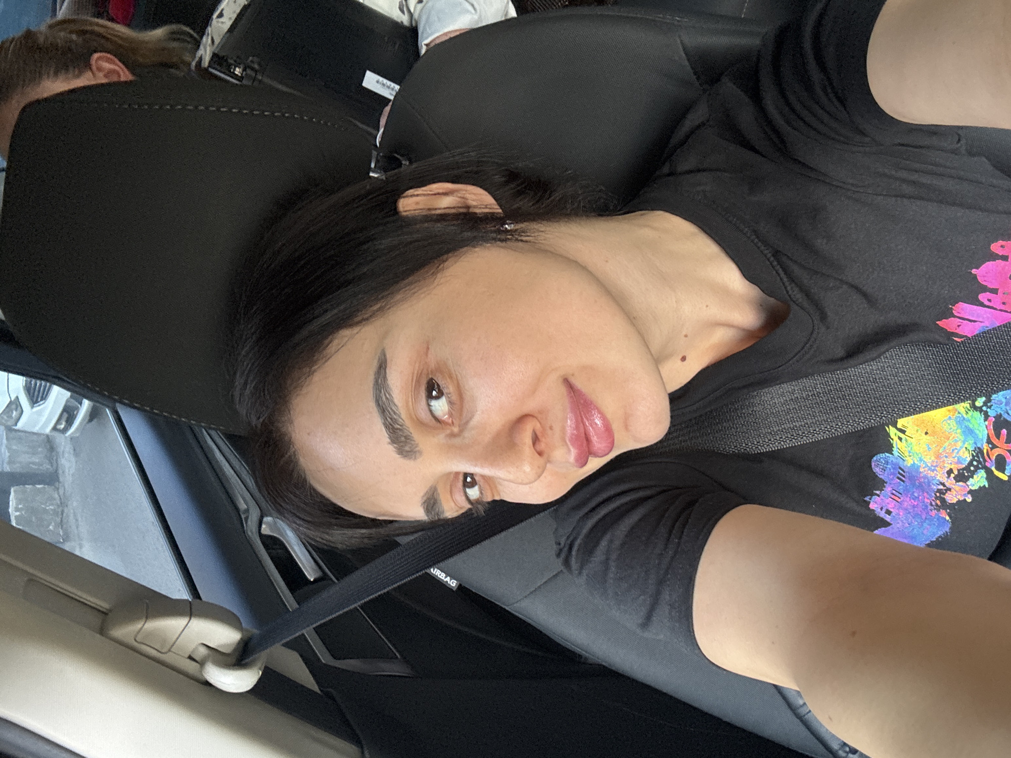 The image shows a woman in a car wearing a black t-shirt with a colorful design and a seatbelt. The location appears to be Tbilisi, Georgia, based on the view out the car window, though specific landmarks are not visible.