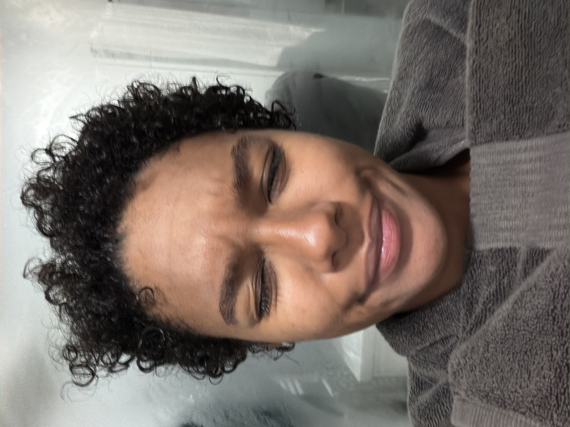 A close-up, angled portrait shows a person with closed eyes, dark curly hair, and brown skin wearing a gray robe. The background is indistinct but appears to be a bathroom interior. The person's facial expression suggests discomfort or strain. The location of the scene is not discernible from the image.