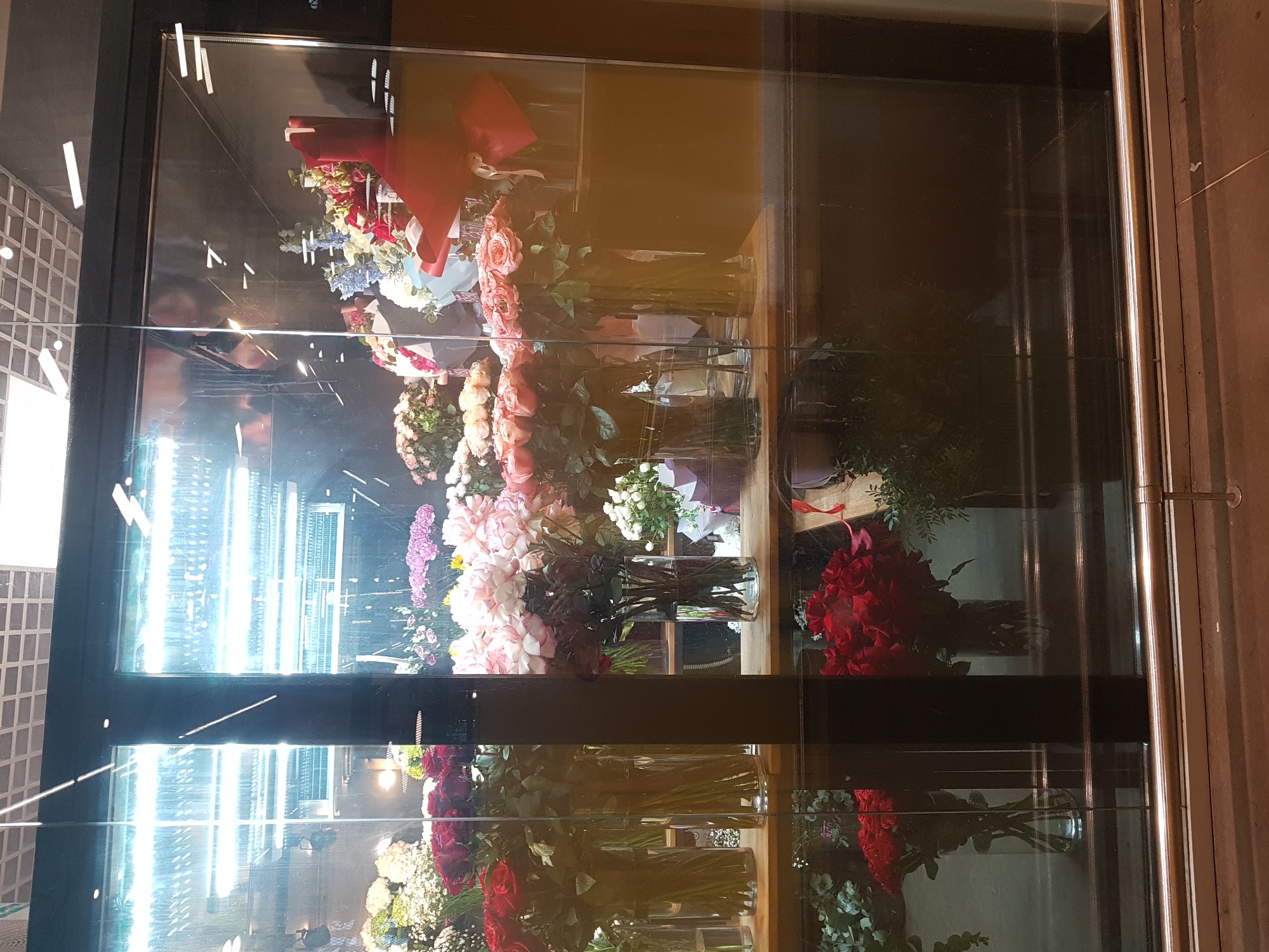 The image depicts a floral display within a glass-fronted refrigeration unit. A variety of flower arrangements, including roses in various colors, are arranged in glass vases. The arrangements are placed on wooden shelves within the refrigerated display. There are red bows visible on some bouquets. The scene is mirrored in the glass. A grid-like surface is visible on the left side through the glass. The image was likely taken indoors. The location is potentially in Saint Petersburg, Russia.