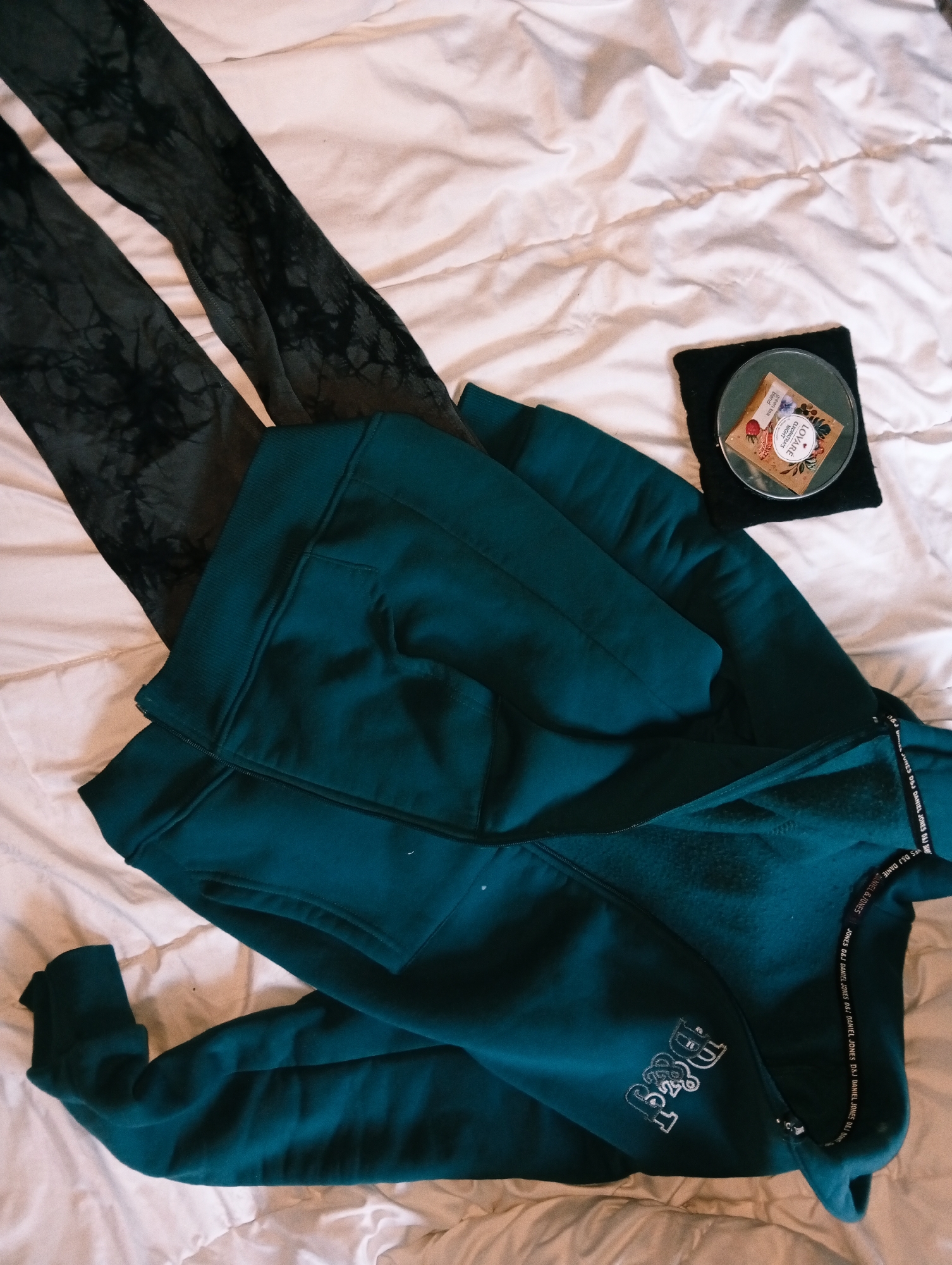 The image shows a flat lay of clothing and other items on a white, crumpled duvet cover, presumably on a bed. To the left are a pair of leggings with a dark tie-dye or marbled pattern in shades of gray and black. Adjacent to the leggings is a folded dark teal or turquoise hooded sweatshirt. The sweatshirt appears to have a zipper closure and a pocket visible on the upper left section. In the lower right area of the sweatshirt, there is white lettering, possibly a logo or brand name, that appears to read "D&J." Additionally, there is a strip of fabric around the hood with the repetitive text: "JONES D&J DANIEL JONES." To the upper right of the sweatshirt, there is a small black square upon which sits a transparent coaster with a tea bag on it. The tea bag is brown with the name of the tea "LOVARE CLEOPATRA'S NIGHT" printed on it. There is no indication in the image that this is from the city of Poltava, Ukraine.