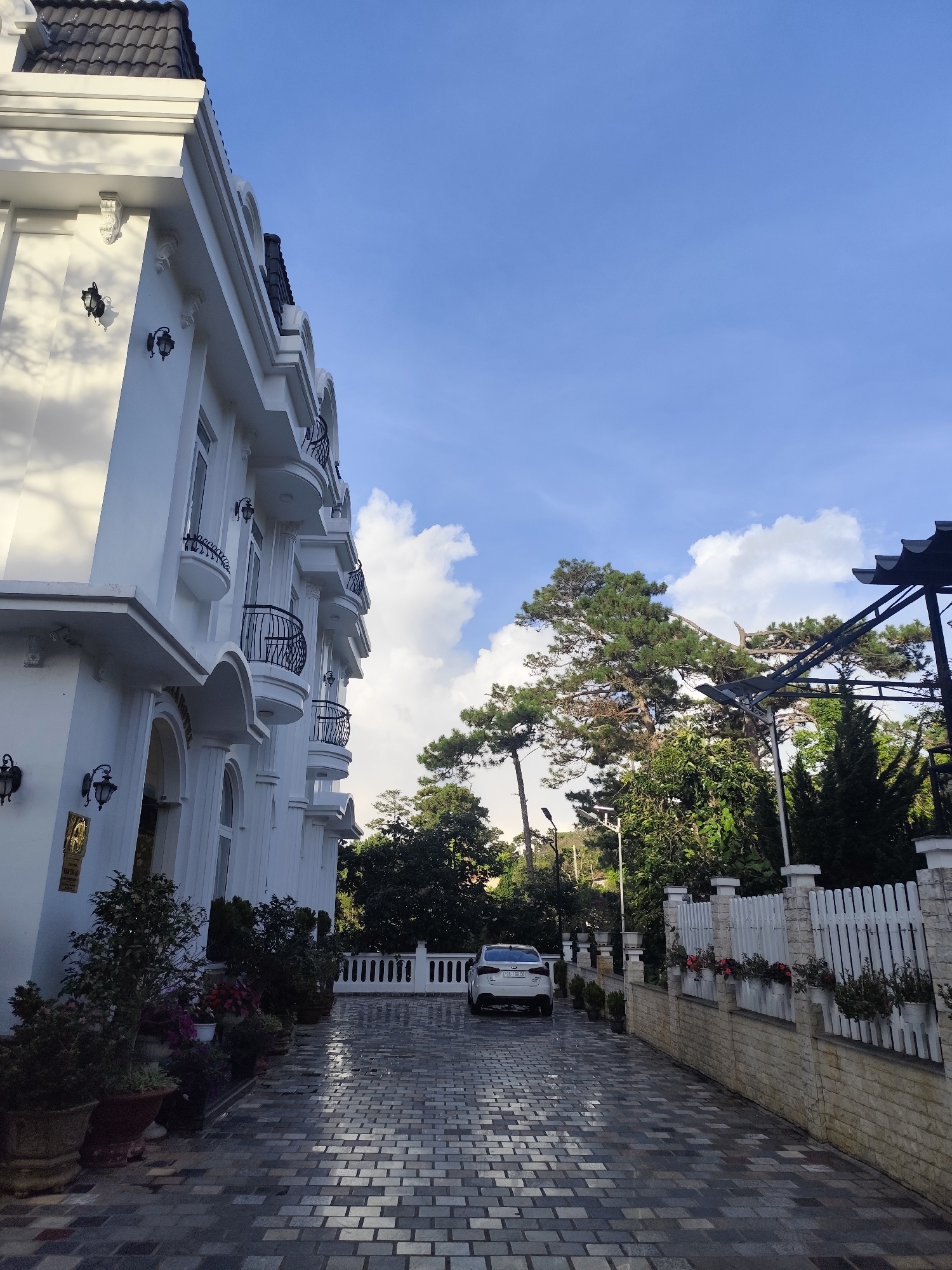 A white, multi-story building with black roof tiles and black wrought-iron balconies is situated on the left side of the image. The building features arched doorways and windows. A white car is parked on a paved driveway in front of the building. The driveway is made of grey and black rectangular pavers. A white picket fence runs along the right side of the driveway, behind which are trees and a glimpse of a metal structure. The sky is blue with scattered white clouds. The scene appears to be in Da Lat, Vietnam, known for its European-inspired architecture and pine trees. The presence of the car and the well-maintained building and grounds suggest a residential or hotel setting. The lighting indicates it is daytime, with sunlight casting shadows.