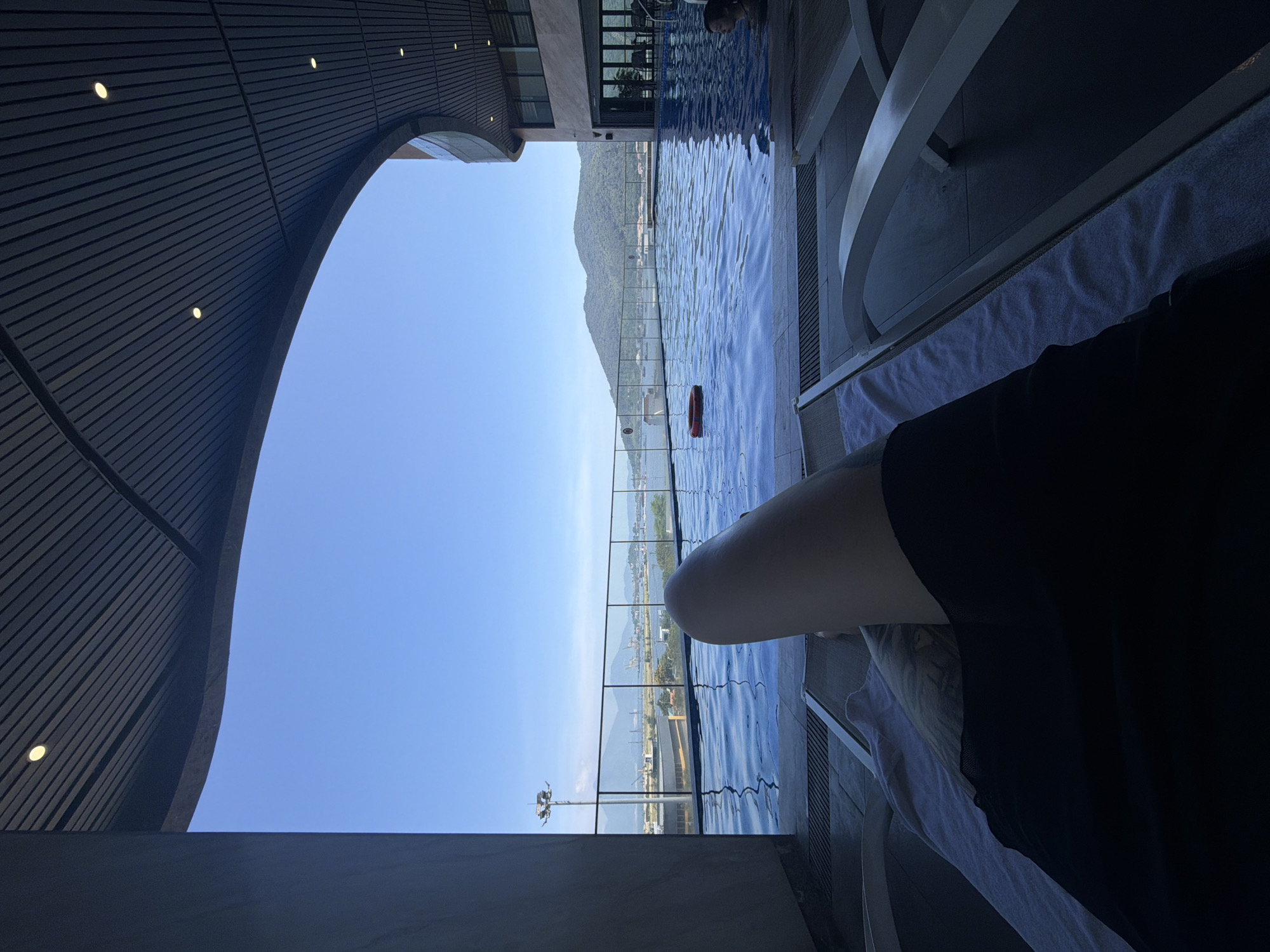 The image captures a serene and luxurious resort scene, likely in Đà Nẵng, Vietnam, featuring an infinity pool with a panoramic view. In the lower right foreground, a bare leg, possibly belonging to someone relaxing on a lounge chair, is visible, alongside dark clothing and a white towel.
The main subject is a blue infinity pool, which occupies a significant portion of the mid-ground. The water is rippled, reflecting the bright sky. A red, circular life preserver floats in the pool. Towards the upper left edge of the pool, a person's head is subtly visible, suggesting someone swimming or enjoying the water.
Beyond the pool's edge, a breathtaking landscape unfolds under a clear, bright blue sky. Large, green-clad mountains dominate the horizon, with a body of water (likely the sea or a bay) nestled between the mountains and the foreground. Along the coastline, some urban development, including buildings and possibly port infrastructure, can be seen.
To the left, a modern architectural element is prominent: a dark, slatted, curved ceiling extends overhead, providing shade to the pool area. Recessed lights are integrated into this ceiling structure. The perspective suggests the photo was taken from a shaded, perhaps private, spot by the pool, looking out over the water towards the distant scenery.
The time of day appears to be bright daylight, with clear weather. The scene evokes a sense of relaxation and enjoyment in a beautiful, upscale setting. There is no visible text in the image.