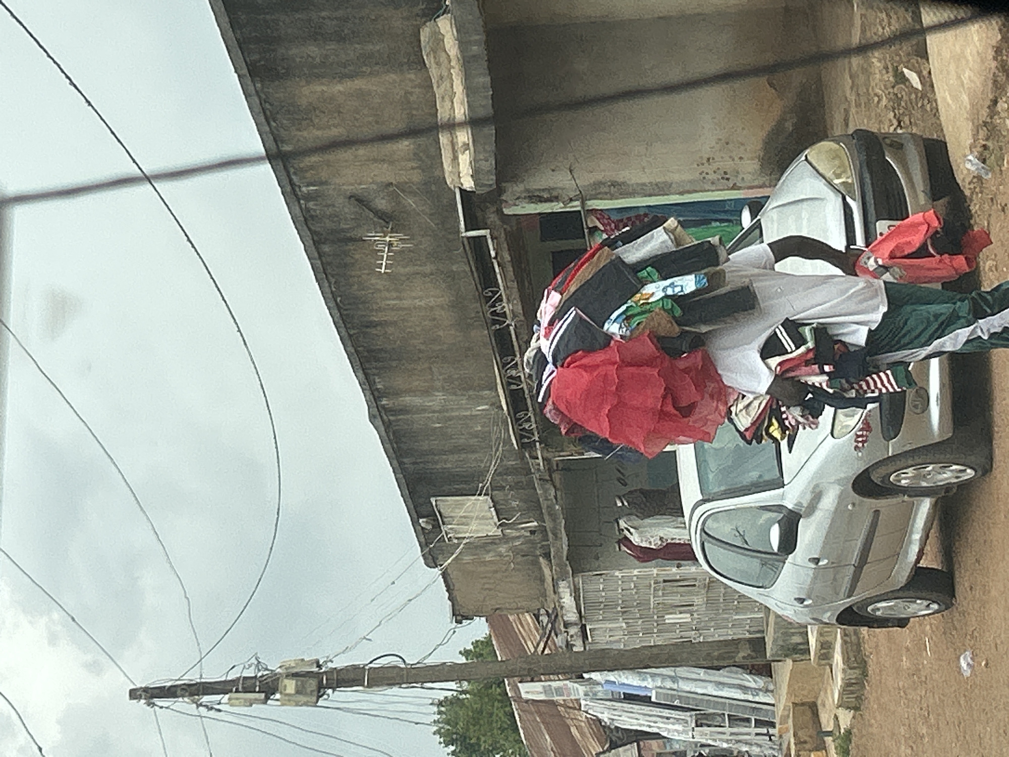 A silver car, overloaded with clothing, is parked beside a building in Jos, Nigeria. The building is weathered, with an antenna on the roof. Power lines stretch across the overcast sky. Disorganized materials are near the building.