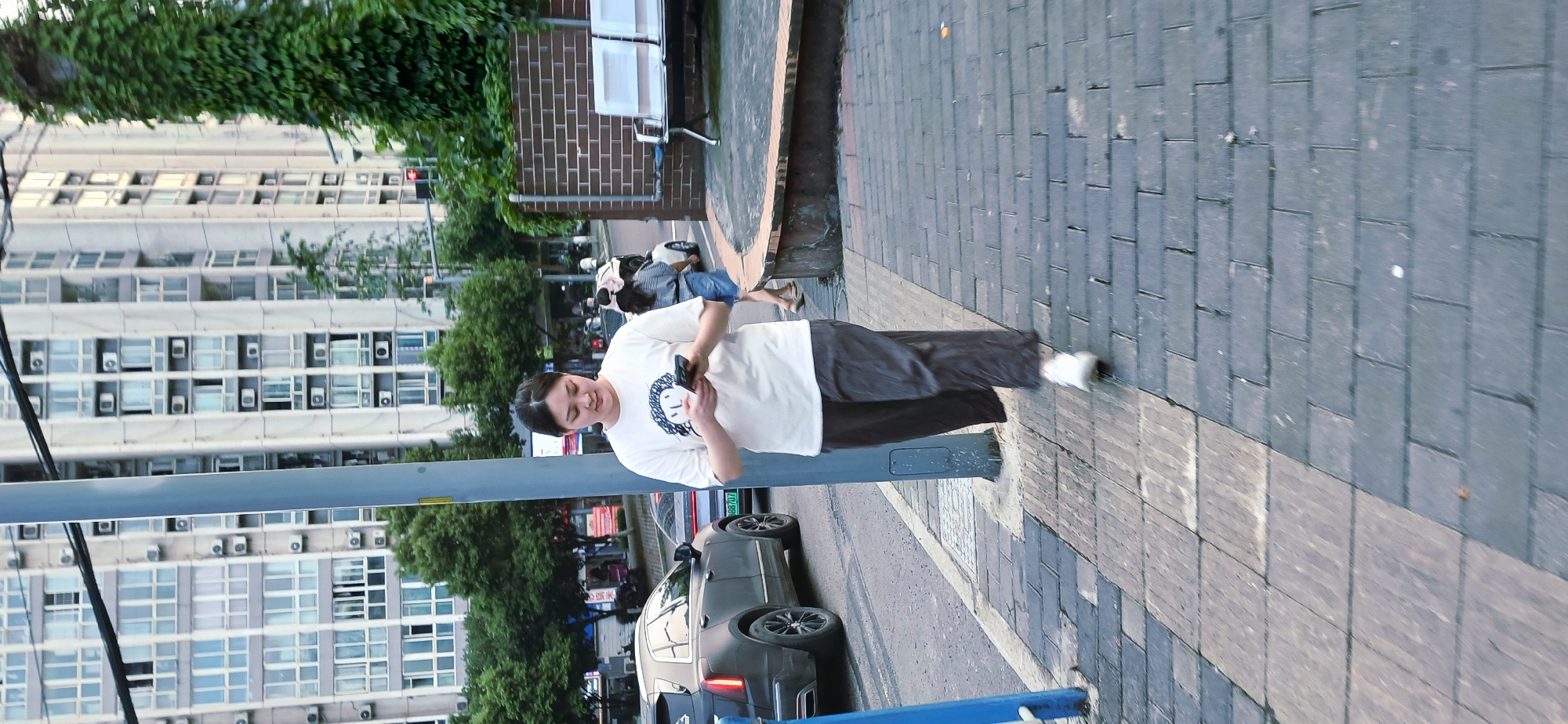 A person stands on a sidewalk in Jiulongpo District, China, looking at their phone. The person is wearing a white t-shirt with a graphic on the front, dark pants, and white sneakers. They are standing next to a gray pole. In the background are a tall building with many windows and air conditioning units, trees, parked cars, and a brick wall. The sidewalk is paved with gray bricks.