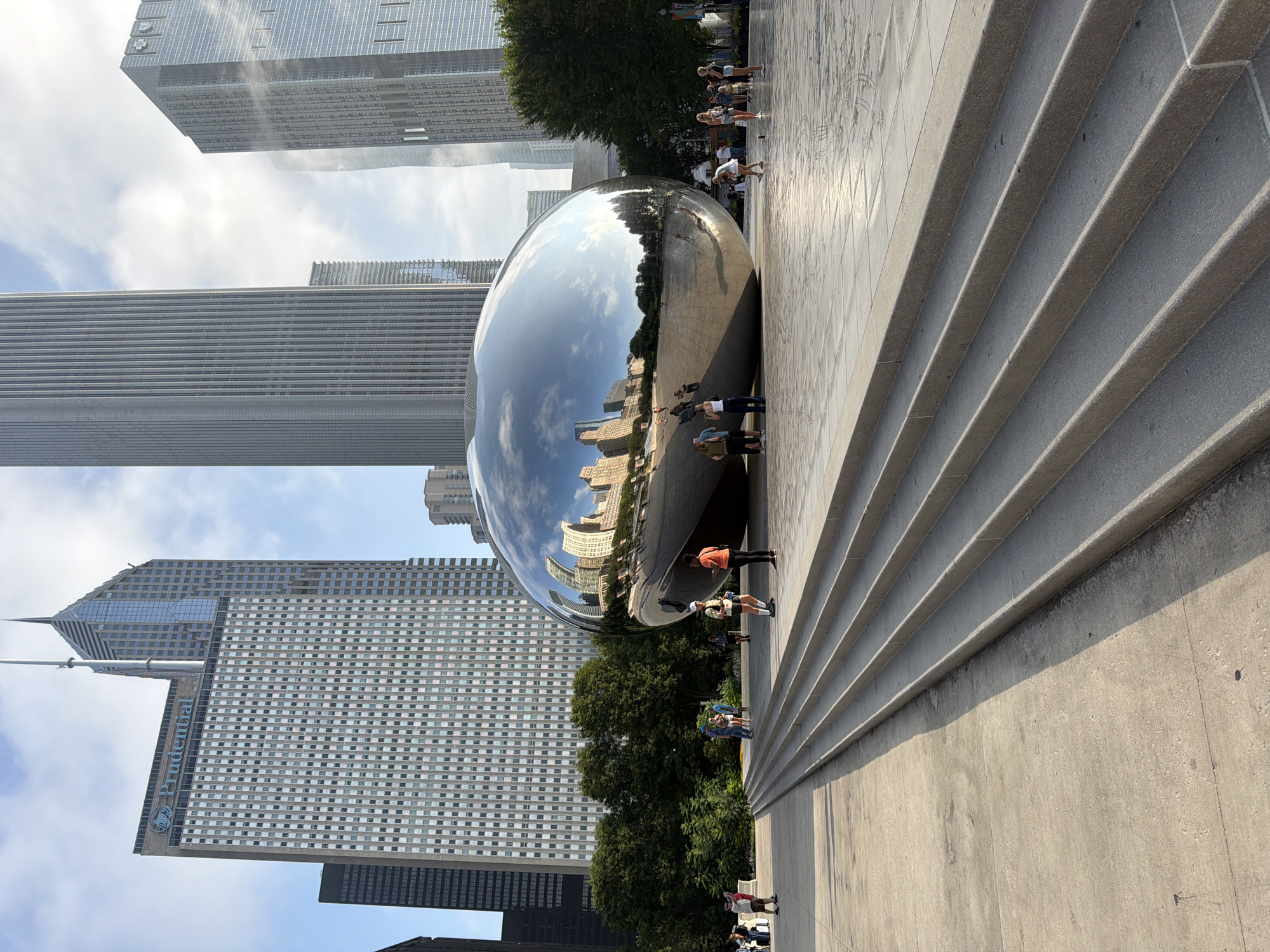 The image depicts "Cloud Gate," a public sculpture by artist Anish Kapoor, commonly known as "The Bean," situated in Millennium Park, Chicago, Illinois, United States of America. The reflective, kidney-shaped sculpture is the central focus, mirroring the surrounding skyscrapers and the sky. In the foreground, a wide concrete plaza features a set of broad, shallow stairs leading towards the sculpture. Several individuals are dispersed across the plaza, some standing and observing the sculpture, others walking. Behind the sculpture, a line of lush green trees separates the plaza from the imposing high-rise buildings that define the Chicago skyline. The sky is partly cloudy, with sunlight casting shadows on the plaza. The architecture visible includes a prominent building with "Prudential" signage.