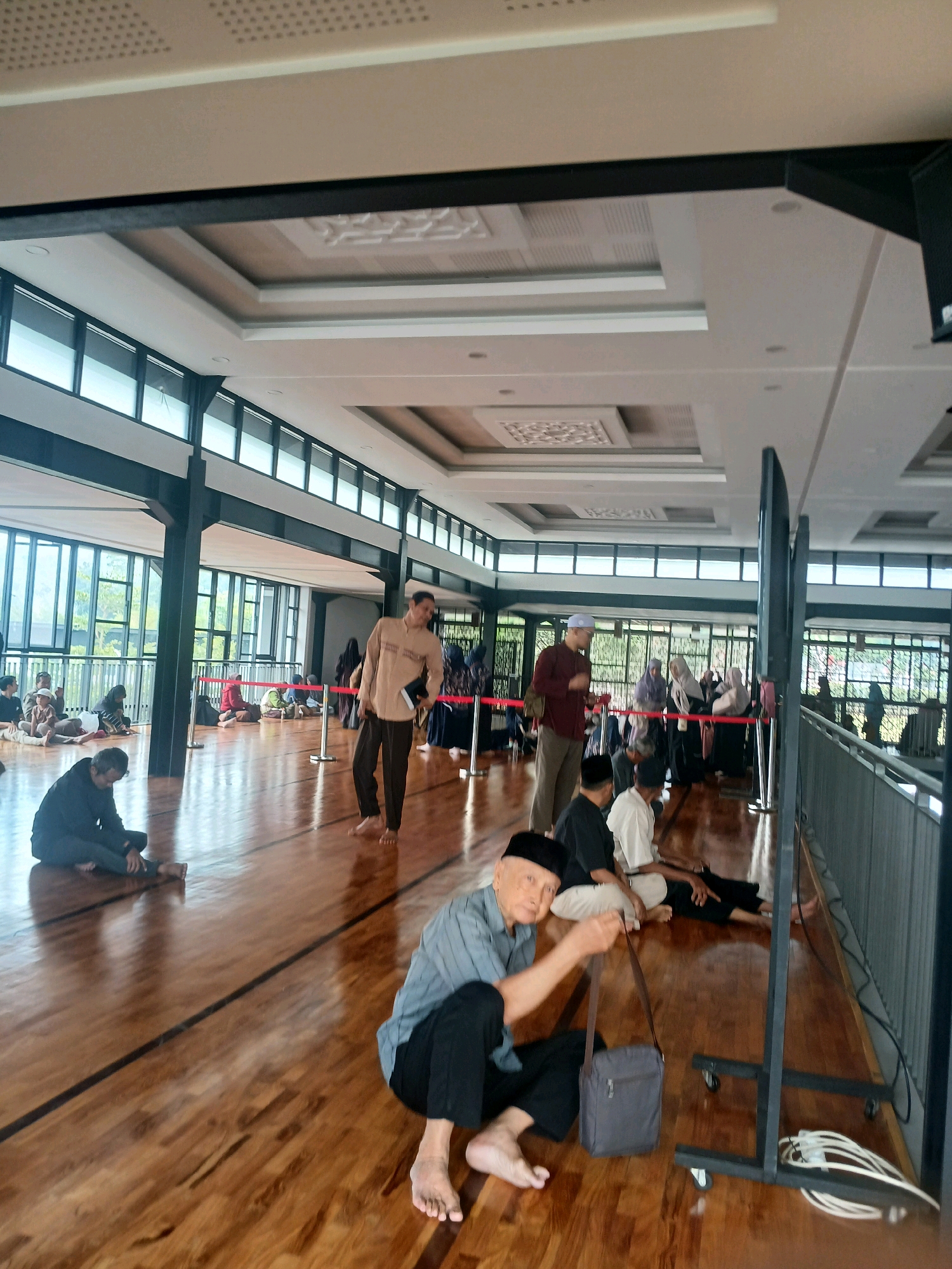 This image depicts the interior of a mosque in Arjasari, Indonesia, likely during a prayer gathering or religious event. The space features a polished wooden floor with dark lines demarcating areas. The ceiling is ornate with recessed lighting and decorative panels. Large windows with black frames line the left side of the room, allowing natural light to enter and offering a view of greenery outside.
Several individuals, predominantly men, are seated or standing on the floor. Many are dressed in traditional Muslim attire, including long shirts, trousers, and skullcaps. Some individuals appear to be praying or listening to a sermon, while others are conversing or resting. A few women are visible in the background, also dressed modestly.
In the foreground, an elderly man in a blue shirt and black trousers is seated, looking towards the camera and holding a small grey shoulder bag. To his right, a dark metal stand with wheels is positioned, holding several white cables. Other people are scattered throughout the room, some sitting in rows, others in smaller groups. Red rope barriers are set up in some areas, possibly to guide the flow of people or separate sections. The overall atmosphere appears to be one of communal gathering and religious observance.
