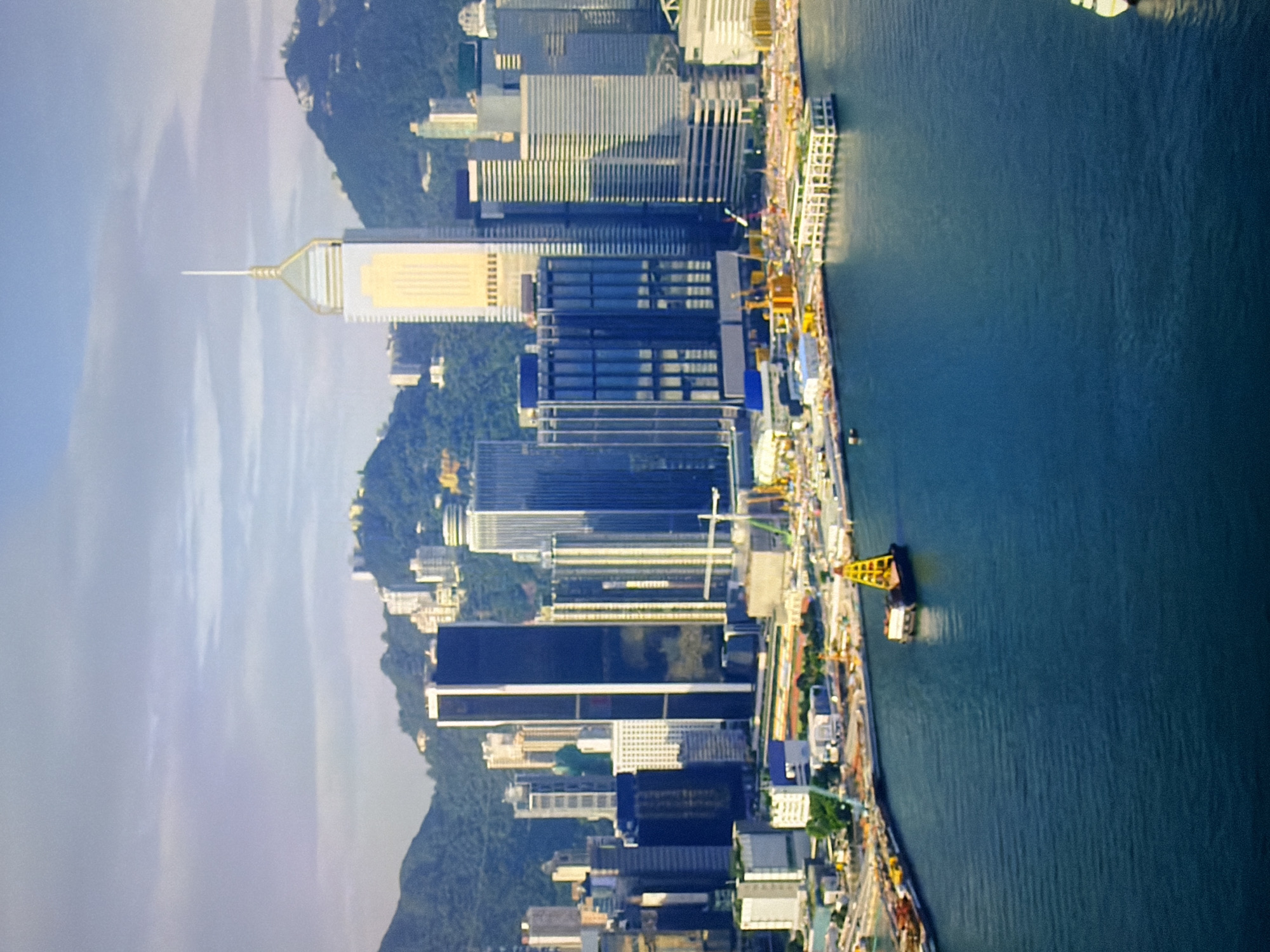 This is a vertical, aerial photograph of a modern city skyline with a large body of water in the foreground. The city features numerous tall skyscrapers, many with distinctive architectural designs. The buildings range in color from dark glass to lighter reflective surfaces, and some are accented with golden or metallic elements. The skyline is backed by lush green, hilly terrain. The body of water appears to be a harbor or bay, with several barges and construction equipment visible along the waterfront, suggesting ongoing development or maintenance. One prominent barge in the water is equipped with a yellow crane structure. The sky is partly cloudy, with a soft, diffused light indicating it might be late afternoon or early morning. The overall impression is one of a bustling, developed urban center integrated with its natural landscape. The city depicted is Hong Kong, not Kyiv, Ukraine.