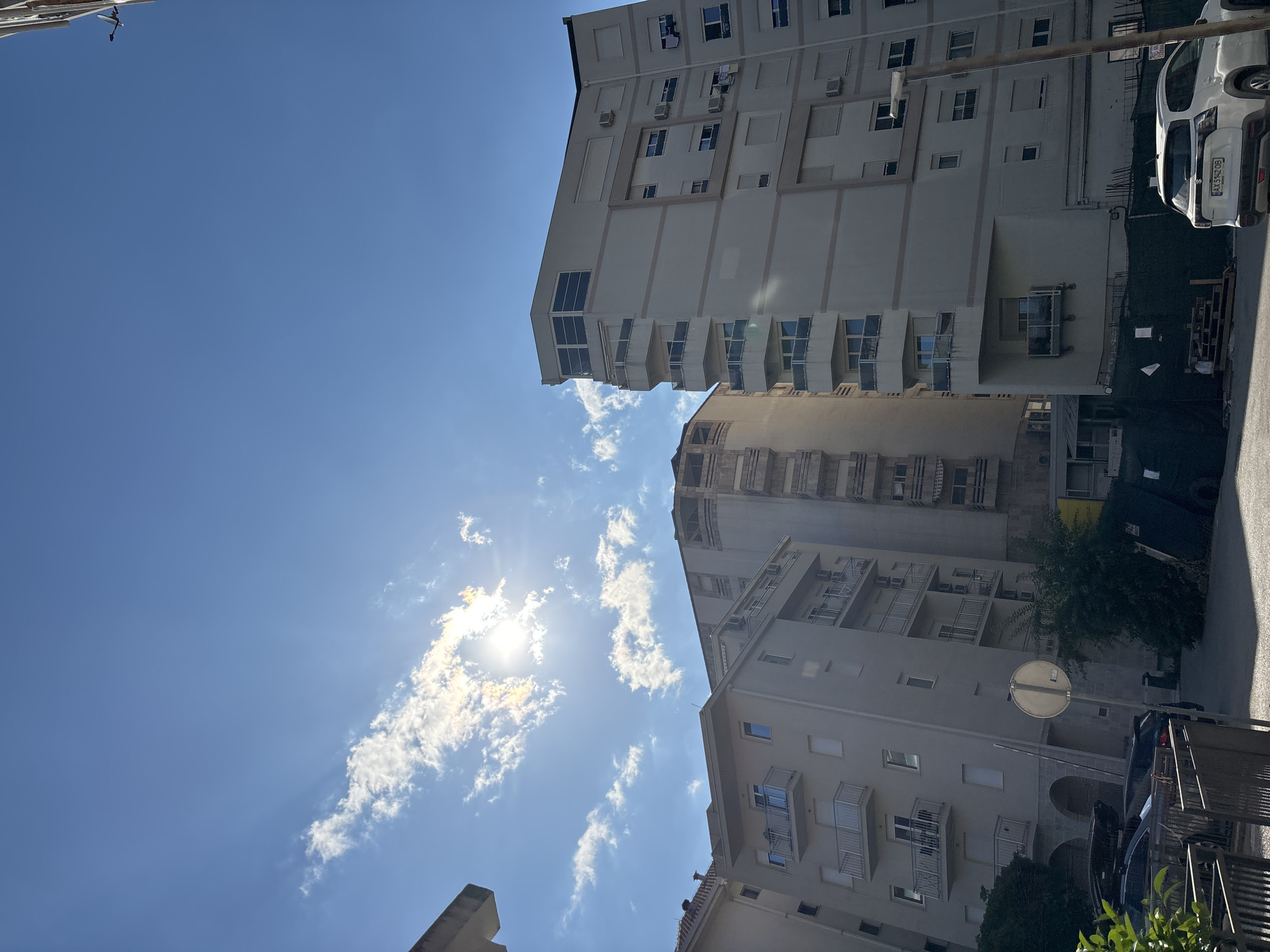 The image shows a street scene in Budva, Montenegro, dominated by tall buildings under a clear blue sky. The sun is bright, casting rays through scattered white clouds, suggesting it is midday. The perspective is looking upwards, giving a sense of the scale of the buildings.
The main subjects are the multi-story buildings, which appear to be residential or office blocks. They are light-colored, with numerous windows and balconies. Some balconies have metal railings and air conditioning units are visible on the facades. One building in the foreground has a distinctive rounded corner.
To the right of the buildings, a white car is parked. Its license plate, "AXS 6478", is partially visible. Further to the right, a street and a fence are visible. There is some greenery, including trees and bushes, interspersed with the concrete structures. A circular street sign is mounted on a pole in front of one of the buildings.
The overall impression is one of a bright, sunny day in an urban environment. The orientation of the photo is unusual, with the sky and buildings appearing at an angle. There is no visible activity of people or any specific event taking place; it is a static snapshot of the location. The image is a typical view of a European coastal city.