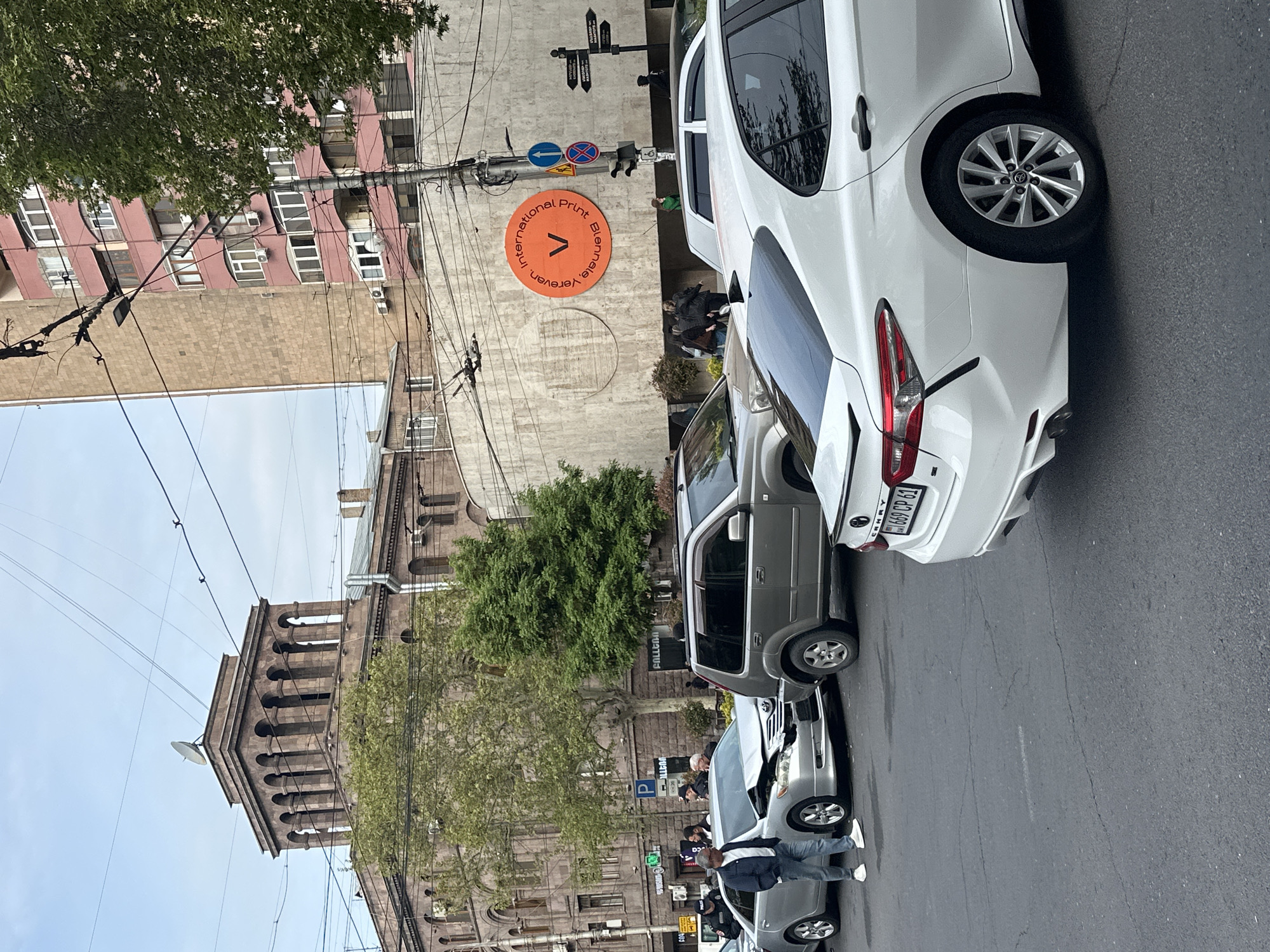 This media file captures an urban street scene during the daytime, likely under a partly cloudy sky. The image is oriented vertically (rotated 90 degrees clockwise), showing a street with parked cars on the right and various buildings and trees on the left.
In the foreground, several cars are visible, including a white SUV/crossover on the far right with its rear visible, a silver SUV/crossover behind it, and a silver sedan closer to the bottom left. A person wearing a dark jacket and jeans is standing next to the silver sedan. Other people are seen walking or standing further in the background.
Architecturally, the scene features a large, ornate stone building with arched window structures, resembling a historic church or government building, on the far left. A smaller satellite dish is mounted on one of its towers. Adjacent to it, a multi-story building with reddish-brown walls and white-framed windows is prominent. Numerous utility poles and overhead power lines crisscross the sky throughout the image. Green foliage from several trees adds natural elements to the urban landscape.
Notable details include an orange circular sign mounted on a building in the mid-ground, displaying the text "International Print Biennale, Yerevan" along with a large letter "V". This strongly suggests the location is Yerevan, the capital of Armenia. Nearby, a blue circular traffic sign with a left-pointing arrow and a red circular sign with a diagonal line are visible, along with a street signpost featuring multiple directional signs. A blue parking sign with a white "P" is also seen.
Visible text includes the aforementioned orange sign. License plates are discernible on the white car ("669 CP 61") and partially on the silver sedan in the foreground ("56 HZ 67"). Part of a sign that reads "GALLERIA" is also visible on a building. The street itself is asphalt, showing some cracks.