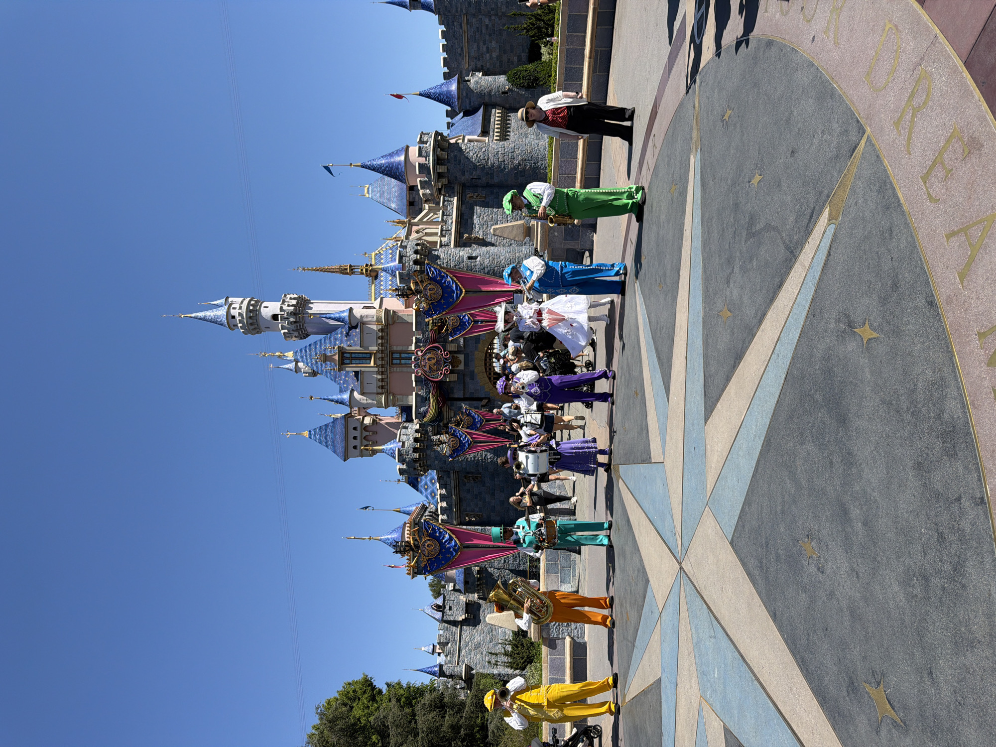 This image captures a daytime scene at Disneyland Park in Anaheim, California, with Sleeping Beauty Castle serving as the prominent backdrop. In the foreground, a group of seven performers dressed in colorful, coordinated costumes are arranged on a large, star-shaped pavement design. The performers are playing various brass and percussion instruments, suggesting a marching band or musical ensemble.
From left to right, the performers are:
1. A person in a bright yellow vest and matching pants, playing a trumpet.
2. A person in an orange vest and matching pants, holding a tuba.
3. A person in a teal vest and matching pants, playing a drum.
4. A person in a purple dress and white hat, holding a cymbal.
5. A person in a purple vest and matching pants, playing a clarinet.
6. A person in a green vest and matching pants, playing a saxophone.
7. A person in a white shirt, red polka-dot vest, and black pants, wearing a straw hat and observing the scene.
Behind the performers, the iconic gray stone structure of Sleeping Beauty Castle is visible, adorned with blue turrets and golden spires. Banners with "70" in large numerals and Mickey Mouse silhouettes are hanging from the castle. The sky is a clear, bright blue. Several other park visitors are visible in the middle ground, walking and standing, some blurred due to motion. The overall atmosphere suggests a festive event or performance taking place in the park.