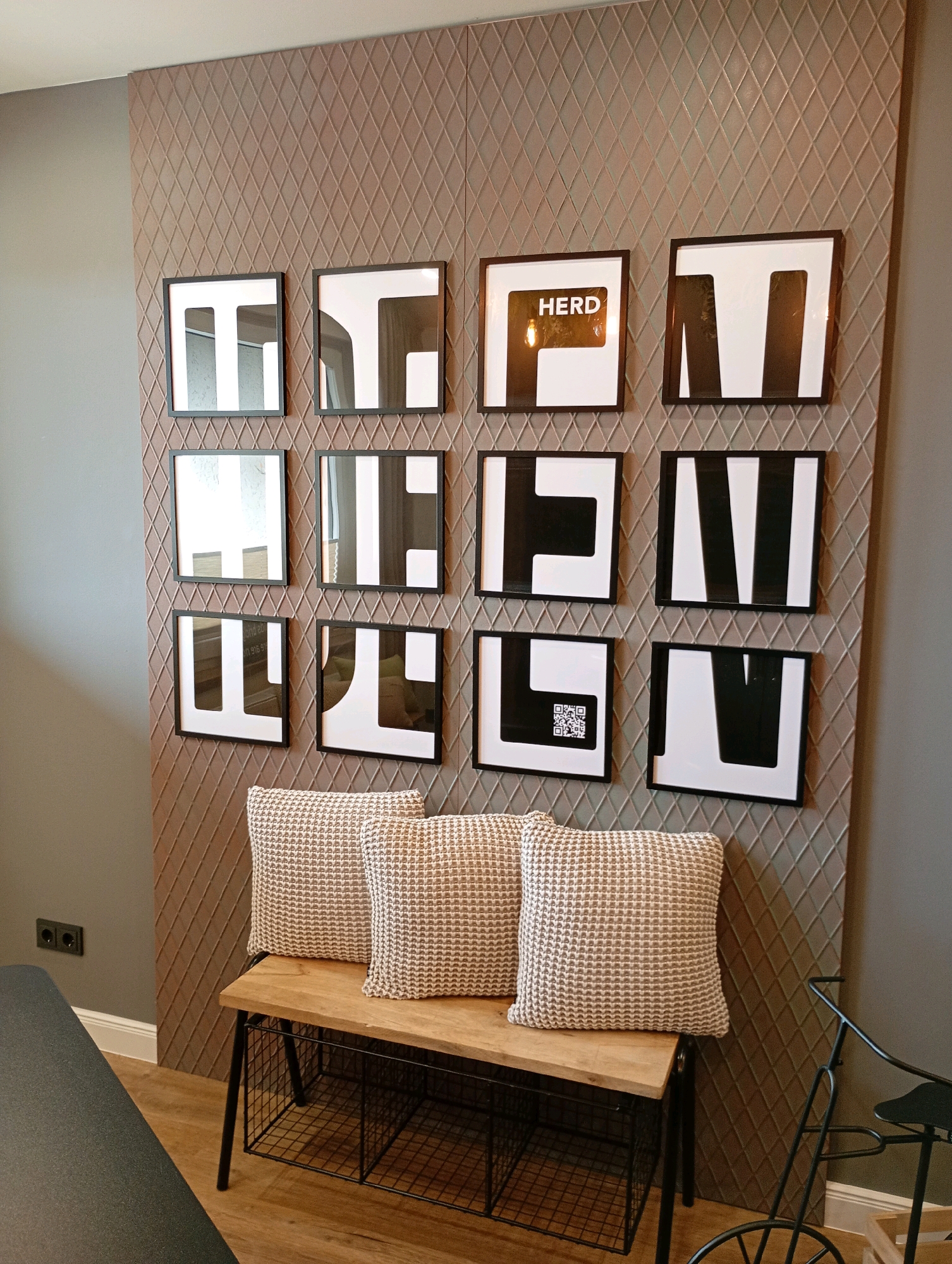 This image depicts an interior scene, likely a living room or a commercial space, featuring a decorative wall with a diamond pattern. Mounted on this wall are twelve framed pieces of modern art, arranged in a 4x3 grid. The artwork primarily consists of abstract geometric designs in black and white, with one piece displaying the word "HERD" within a black frame and a light bulb.
In front of the artwork, there is a wooden bench with a black metal frame and a wire mesh shelf underneath. Two textured throw pillows with a neutral color scheme are placed on the bench. To the left of the bench, a dark-colored object, possibly a sofa or table, is partially visible. On the left wall, an electrical outlet is visible.
The overall aesthetic is contemporary and minimalist. The scene is well-lit, highlighting the textures and patterns of the decor. The image does not contain any people. Based on the context of the request, this scene is located in Bad Sachsa, Germany, suggesting it might be in a hotel, a public building, or a private residence in the town.