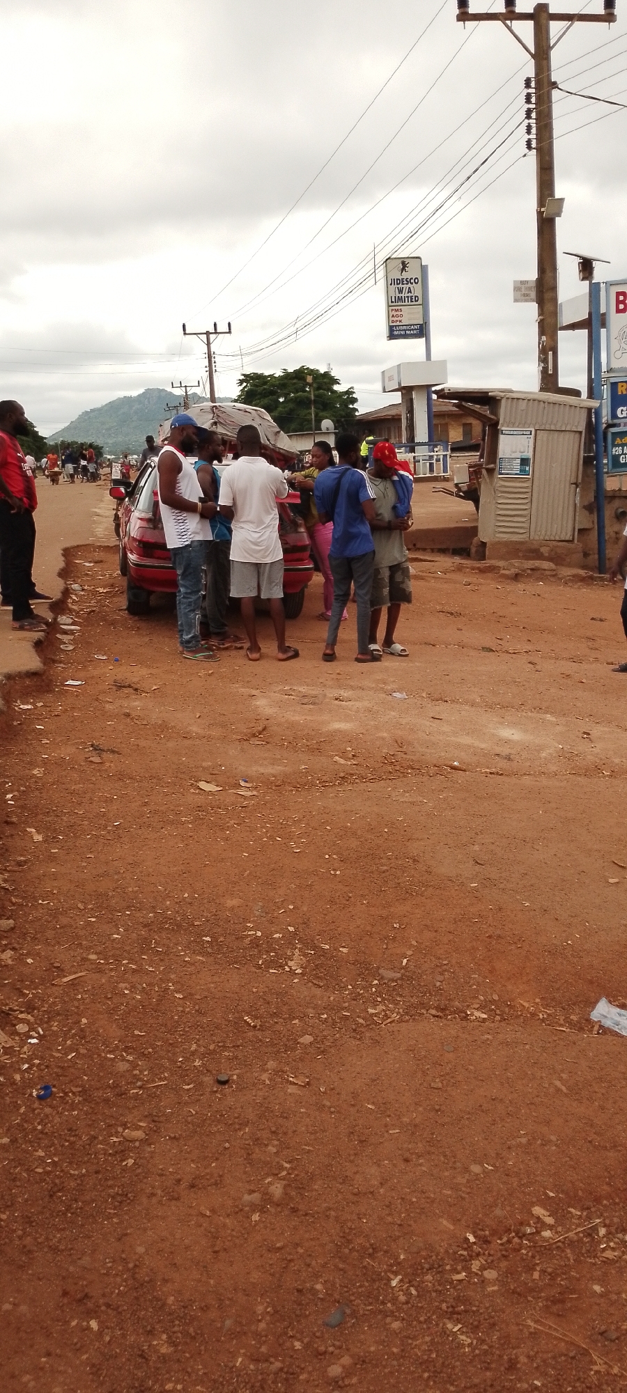 A group of five young Nigerian men are gathered around the rear of a red sedan on an unpaved dirt road. The men are dressed in casual clothing, including t-shirts, shorts, and jeans. One man in a white t-shirt and grey shorts appears to be talking to the others. The man to his left, wearing a blue cap and a white tank top, is looking away. A fourth man, in a white t-shirt, gray shorts, and a red cap, is standing further to the right. Another man in a blue shirt and dark pants is holding something in his arms. The scene appears to be taking place in a small town or rural area. In the background, there are utility poles with power lines, a sign that reads "JIDESCO (W/A) LIMITED," and some low-rise buildings. A large hill or mountain is visible in the distance. The ground is covered in reddish-brown dirt with some scattered debris. The sky is overcast. The image was taken in Gboko, Nigeria.