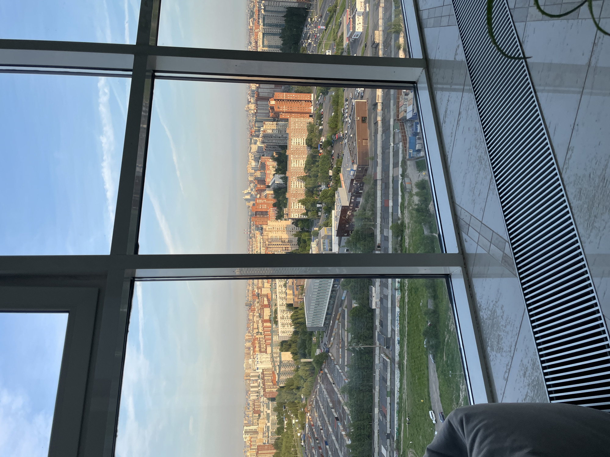This is an indoor, eye-level, vertical shot looking through a large window at a cityscape. The window frame is visible in the foreground, dividing the scene into four panes, with a grey, padded chair on the left. The floor is tiled and reflective, with a silver heating grate running along the bottom. Outside the window, the view is of a dense urban environment with numerous apartment buildings, some of which are orange-brown and others are grey or white. There are also many rows of garages or storage units and some industrial buildings. Trees and green spaces are interspersed throughout the cityscape. A highway with traffic is visible on the right side of the image. The sky is blue with some wispy white clouds. The lighting suggests it is late afternoon or early evening.