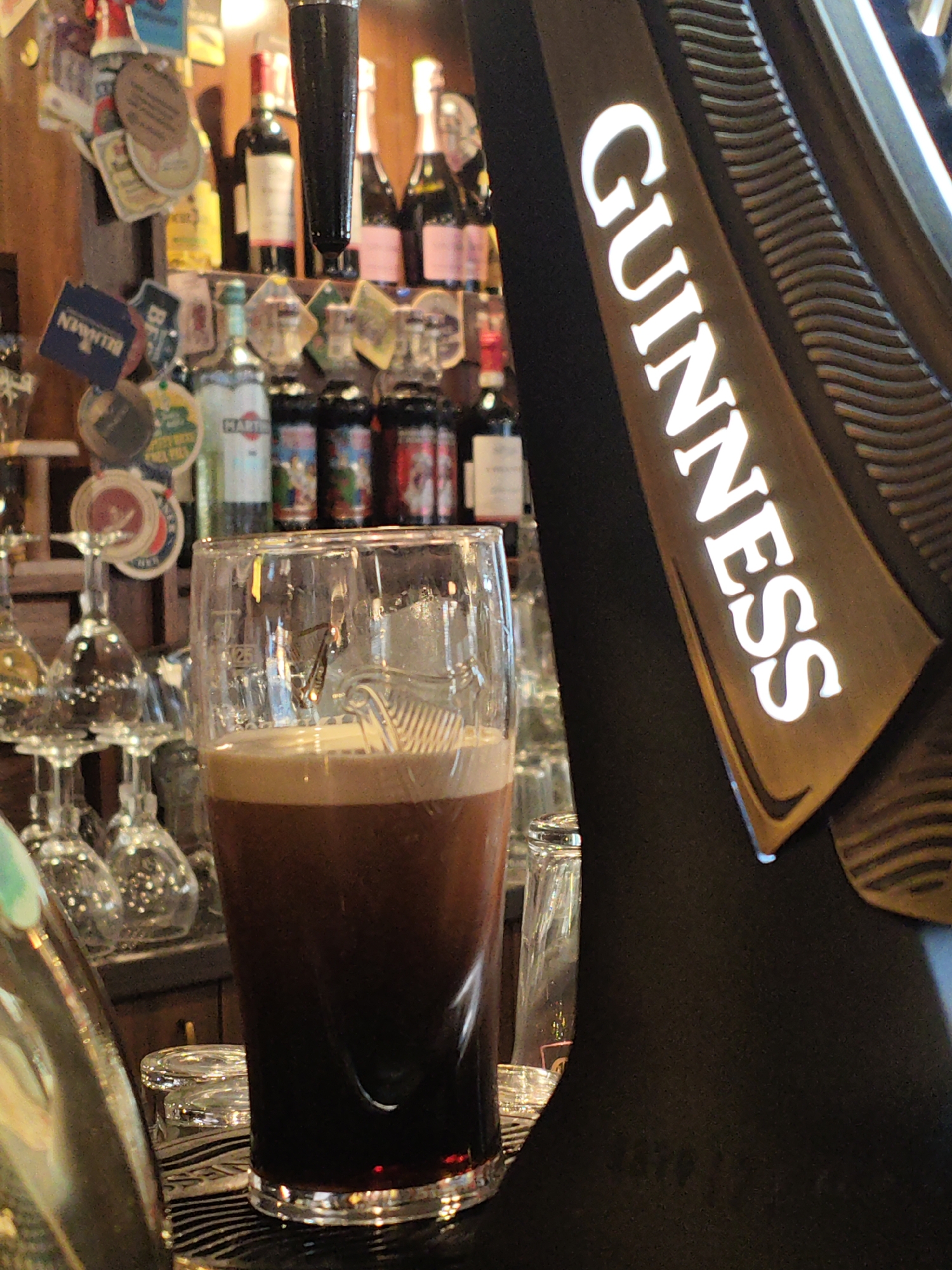 A close-up, eye-level shot captures a pint of Guinness beer on a counter in a dimly lit establishment. The dark beer fills most of a glass, topped with a thick, creamy head of foam. To the right, a prominent Guinness logo illuminates the scene, its white lettering stark against a golden background. In the background, rows of bottles and glassware are visible, suggesting a bar or pub setting. The overall atmosphere is warm and inviting, hinting at a relaxed evening. The establishment is not specified to be in Kyiv, Ukraine.