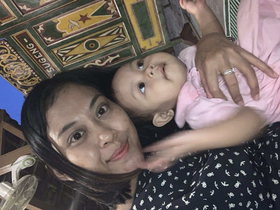 A medium shot depicts an adult female holding an infant. The adult female, with dark, short hair and a light complexion, looks directly at the camera with a slight smile. She wears a dark blue garment with a white pattern and a ring on her left ring finger. She is holding an infant dressed in a light pink collared top, positioned to her right. The infant has dark eyes, a light complexion, and is looking upward and to the right of the frame. In the background, to the left, two ceiling fans are partially visible, along with wooden structural elements. To the right, a richly decorated wooden panel with intricate carvings and painted designs in green, yellow, red, and gold is visible. The word "SUGENG" is legible on a horizontal white strip within this panel, indicating a traditional Javanese architectural style. The scene is located in Mojorejo, Indonesia, under what appears to be ambient or artificial lighting, possibly indoors or in a covered outdoor area.