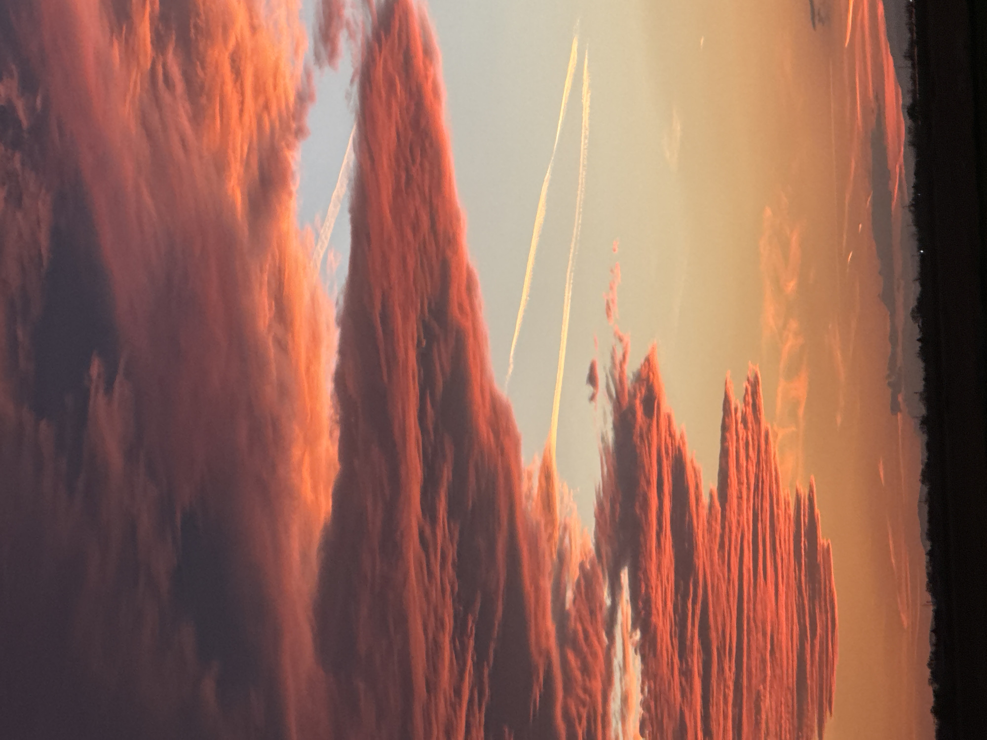 The image shows a low-angle, daytime view of red-tinged, vertical cloud formations resembling mountain peaks in the sky. Contrails are visible in the open sky above the clouds. The lower right of the image shows a blurry, dark edge. There is no evidence of Malenovo, Bulgaria. There are no people or subjects.