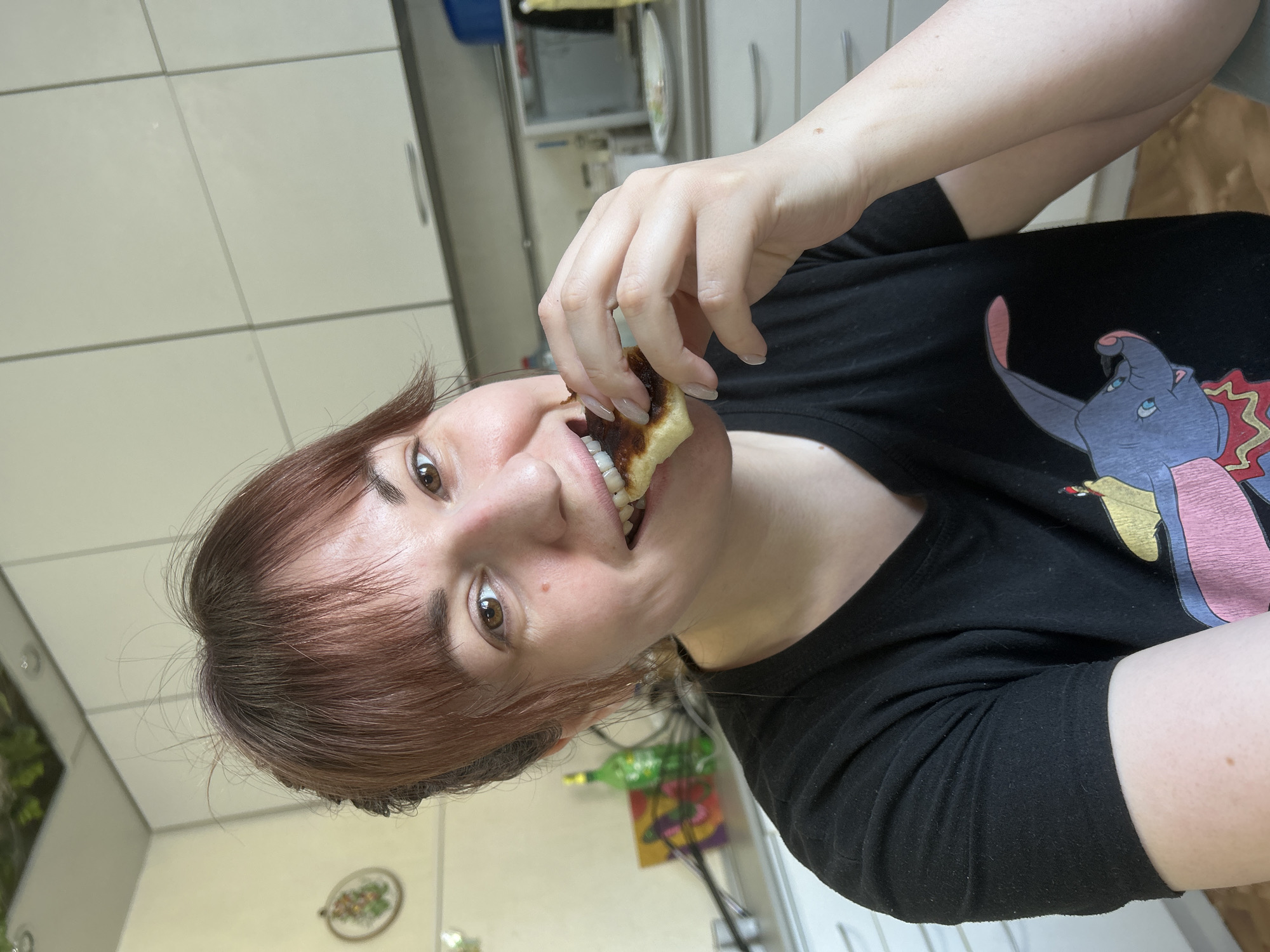 An adult female is depicted indoors, likely in a residential kitchen. The specific city of Фоки, Russia, cannot be determined from the visual evidence. She has medium-toned skin, brown eyes, and shoulder-length reddish-brown hair styled with bangs, pulled back from the sides. She is wearing a black short-sleeved t-shirt featuring a partial graphic of Dumbo the elephant. Her head is angled slightly to her left, and she is looking directly at the camera with a smile, showing her teeth. Her right hand, with light-colored manicured fingernails, holds a piece of food, possibly a pancake or fried pastry with a dark, caramelized, or slightly burnt surface, which she is biting into with her front teeth.
The background consists of light-colored kitchen cabinets with silver handles. A light-colored countertop is visible, upon which various items are placed. To the left, a green glass bottle and a colorful framed picture or artwork are visible on the countertop, with some dark cables behind them. A round, framed decorative item, possibly needlework, hangs on the wall further to the left. To the right, a microwave oven with a reflective door is visible on the countertop, next to a white plate with a patterned rim. The walls are light-colored. The scene is brightly lit, suggesting daylight or strong artificial illumination.