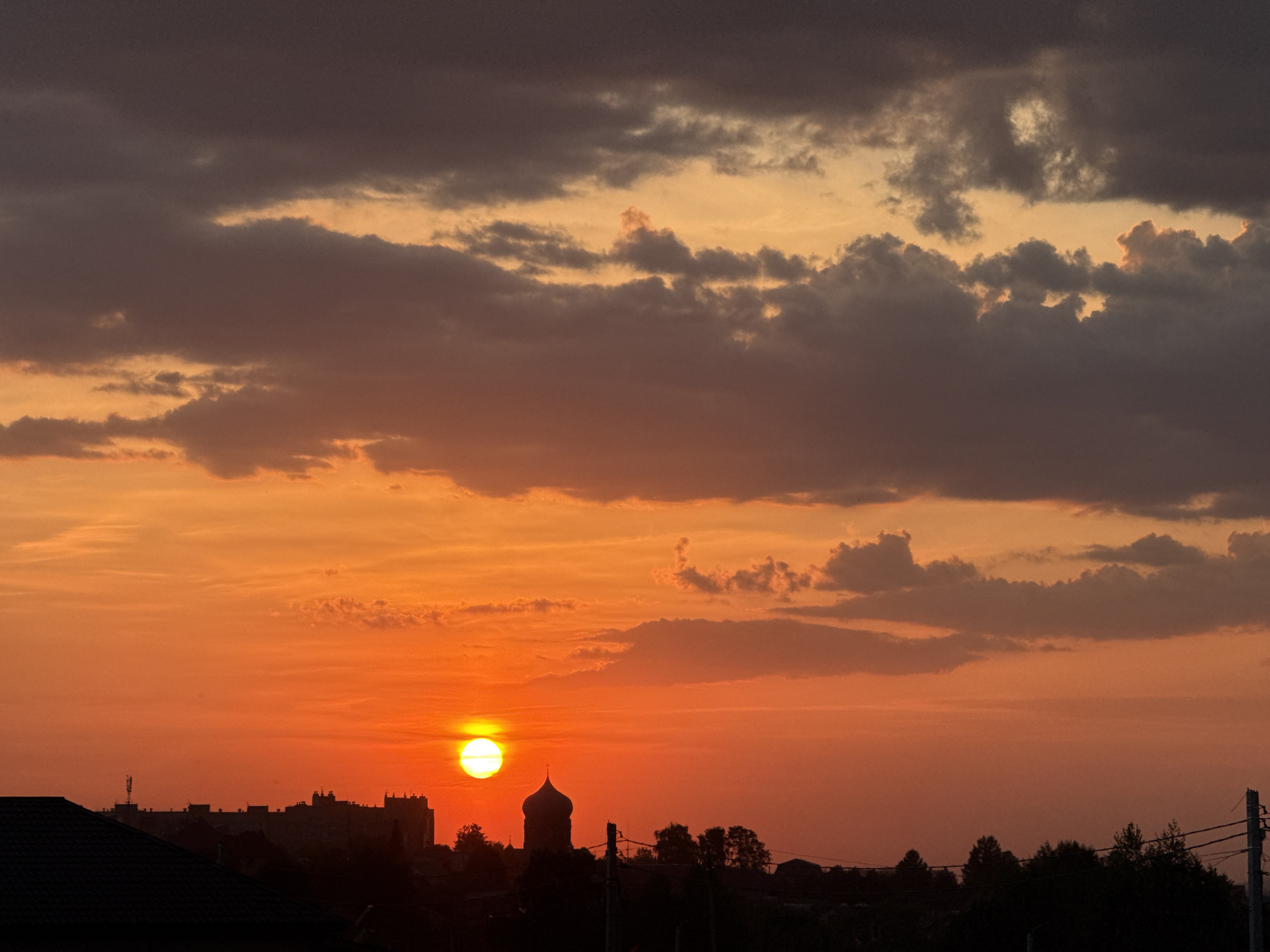 The image depicts a sunset over the city of Lobanovo, Russia. The sky is a gradient of orange, yellow, and purple hues, with clouds scattered throughout. The sun is visible, positioned low on the horizon. In the foreground, there is a silhouette of a building, trees and a church dome. Electrical poles are also present.