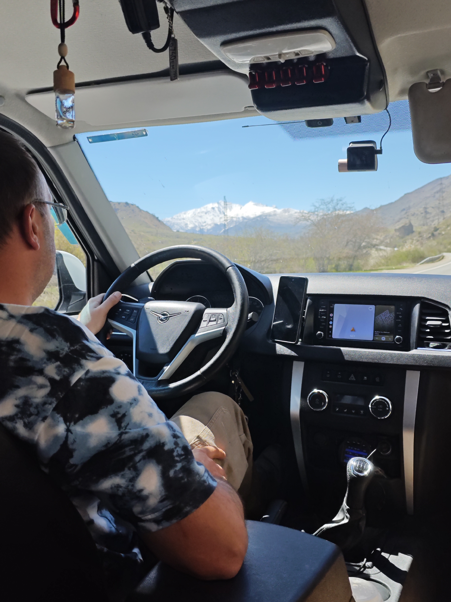 The image shows the interior of a car, specifically the driver's seat and dashboard, with a view out the windshield towards a mountainous landscape. A man is seen from behind, seated in the driver's seat, holding the steering wheel. The dashboard features a central touchscreen displaying a map, a mobile phone mounted to the left of the screen, and various controls below. An air freshener hangs from the rearview mirror. Through the windshield, a mountain range with snow-capped peaks is visible. The road ahead is winding. The location is Bedyk, Russia. The presence of a manual transmission is noted.