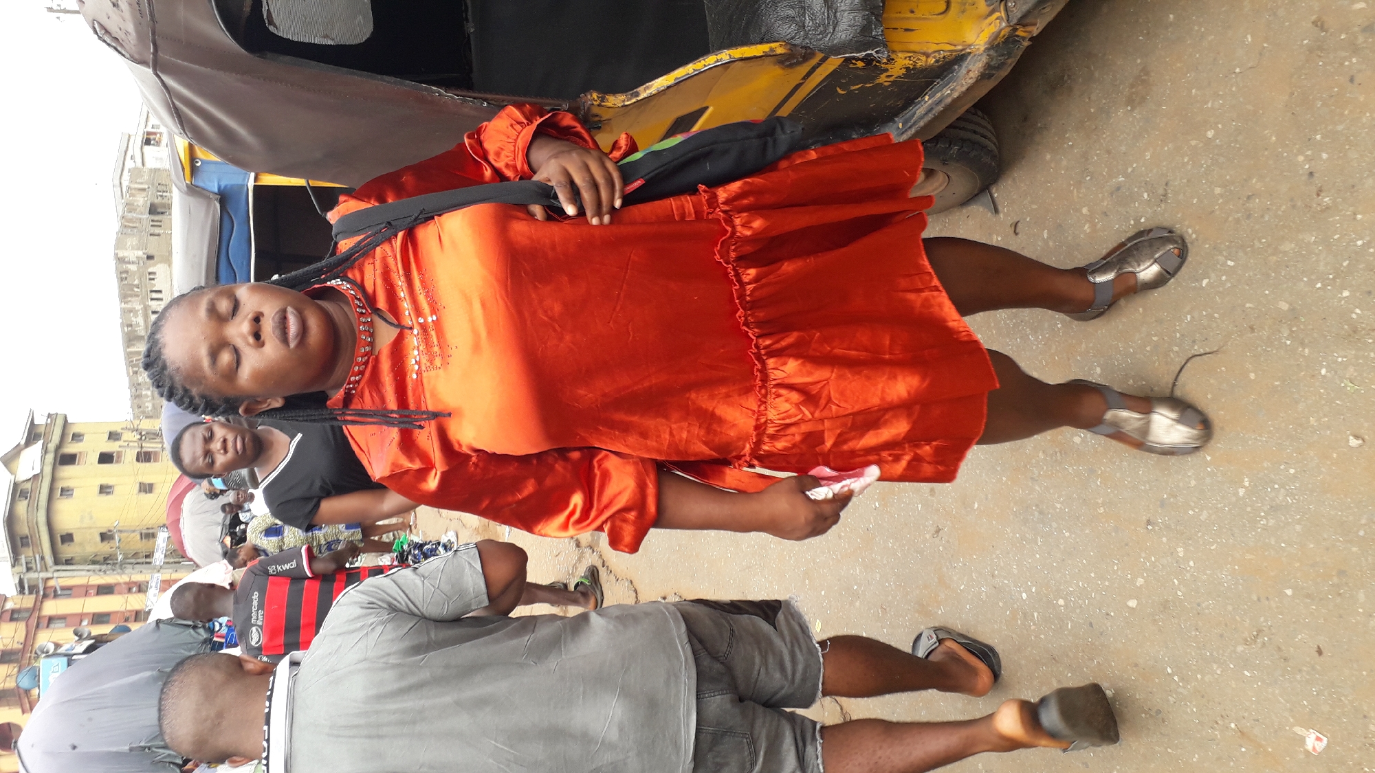 The image shows a woman in Nkpor, Nigeria, wearing an orange dress and silver shoes, standing near a yellow vehicle, possibly an auto rickshaw. She is holding a small white object, perhaps a phone or paper, and has a black bag slung over her shoulder. The background reveals a crowded urban scene with buildings and other people. The pavement beneath the woman's feet is concrete. Other people in casual attire are visible, suggesting a street scene.